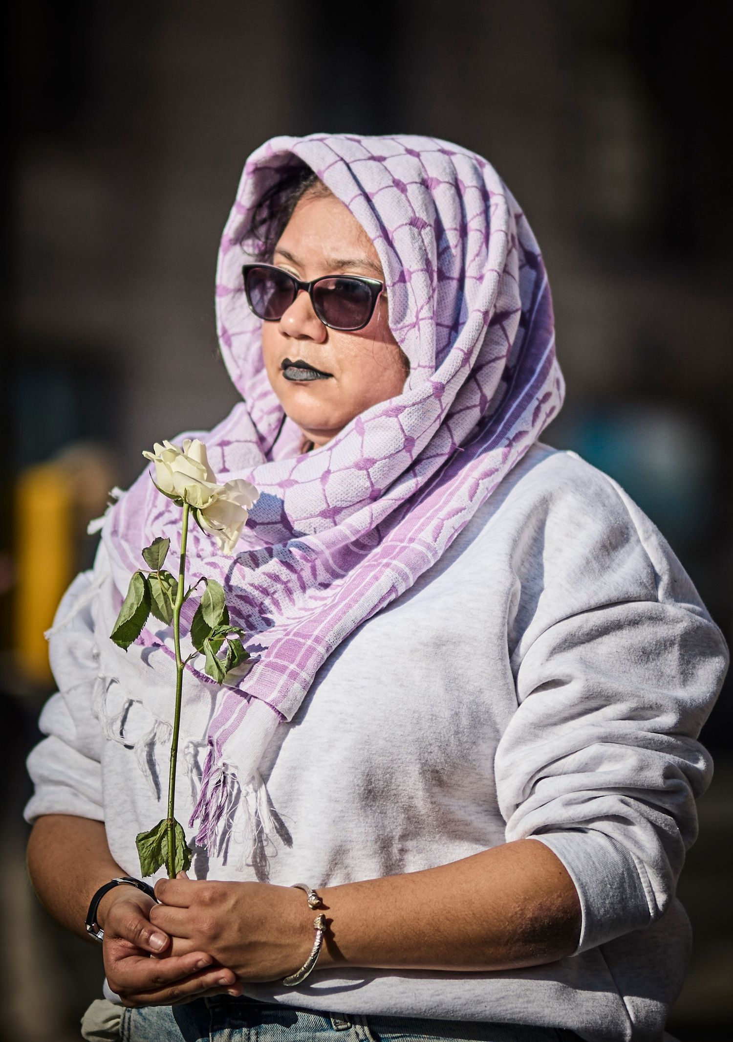 A person in a lilac patterned hijab and sunglasses holding white flowers.