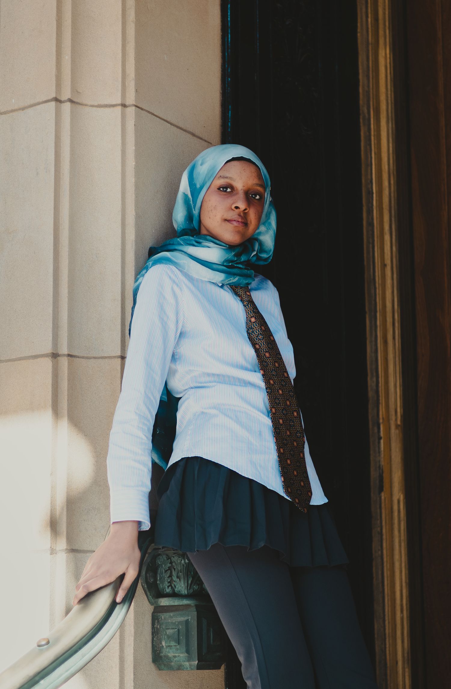 Young woman in blue hijab and tie leans against ornate stone doorway in professional attire.