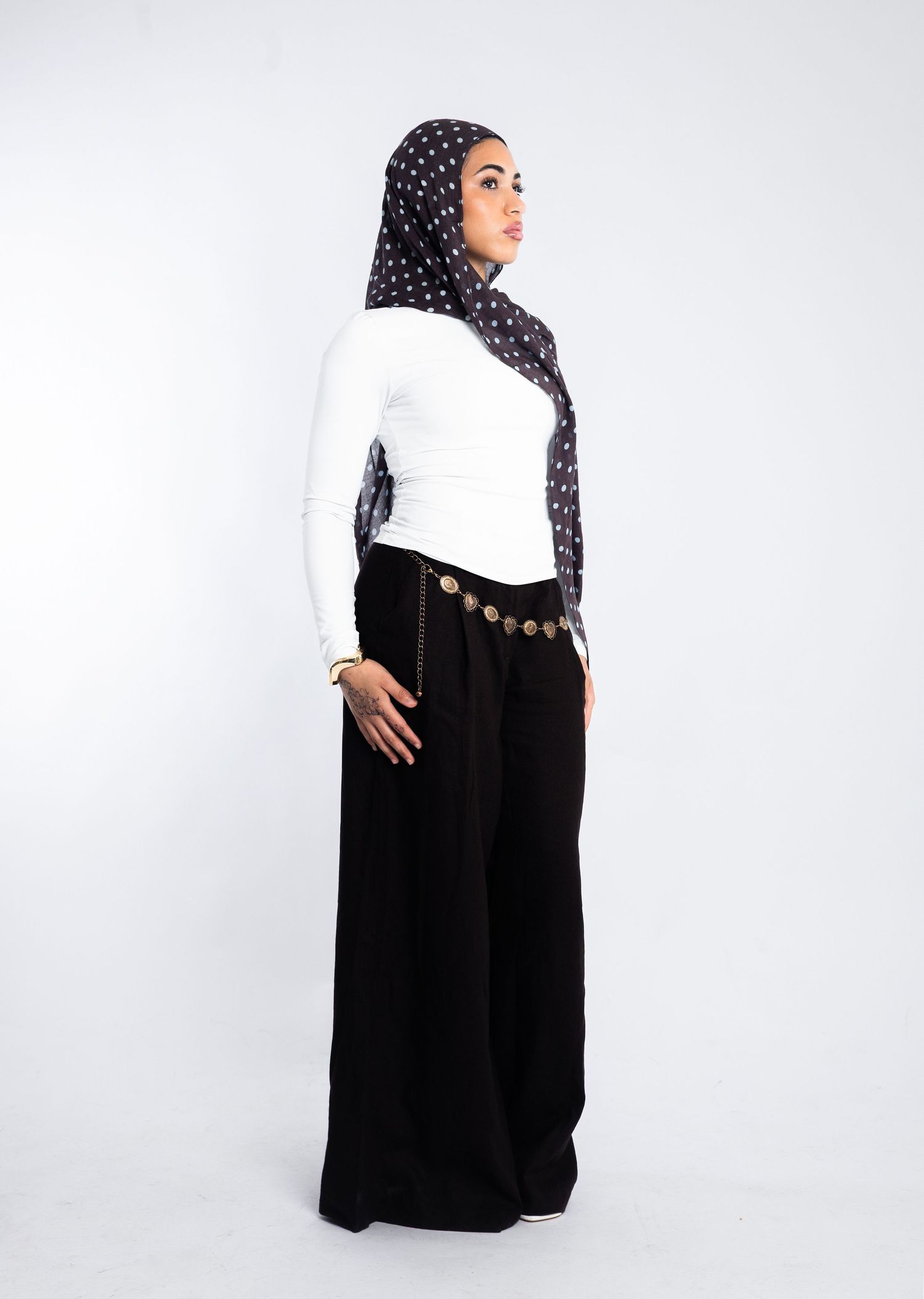 Woman in white long-sleeve top, black wide-leg pants with gold coin belt, and dark polka dot hijab, side profile view.