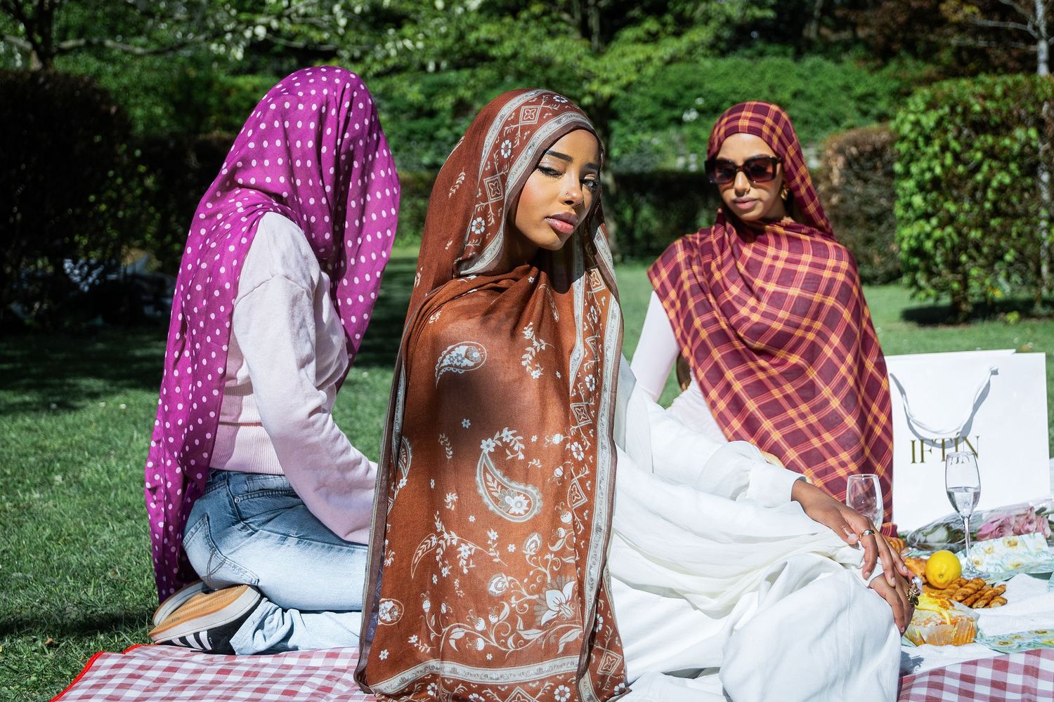 Three women wearing colorful hijabs enjoy an outdoor picnic on a sunny day in a lush green park.