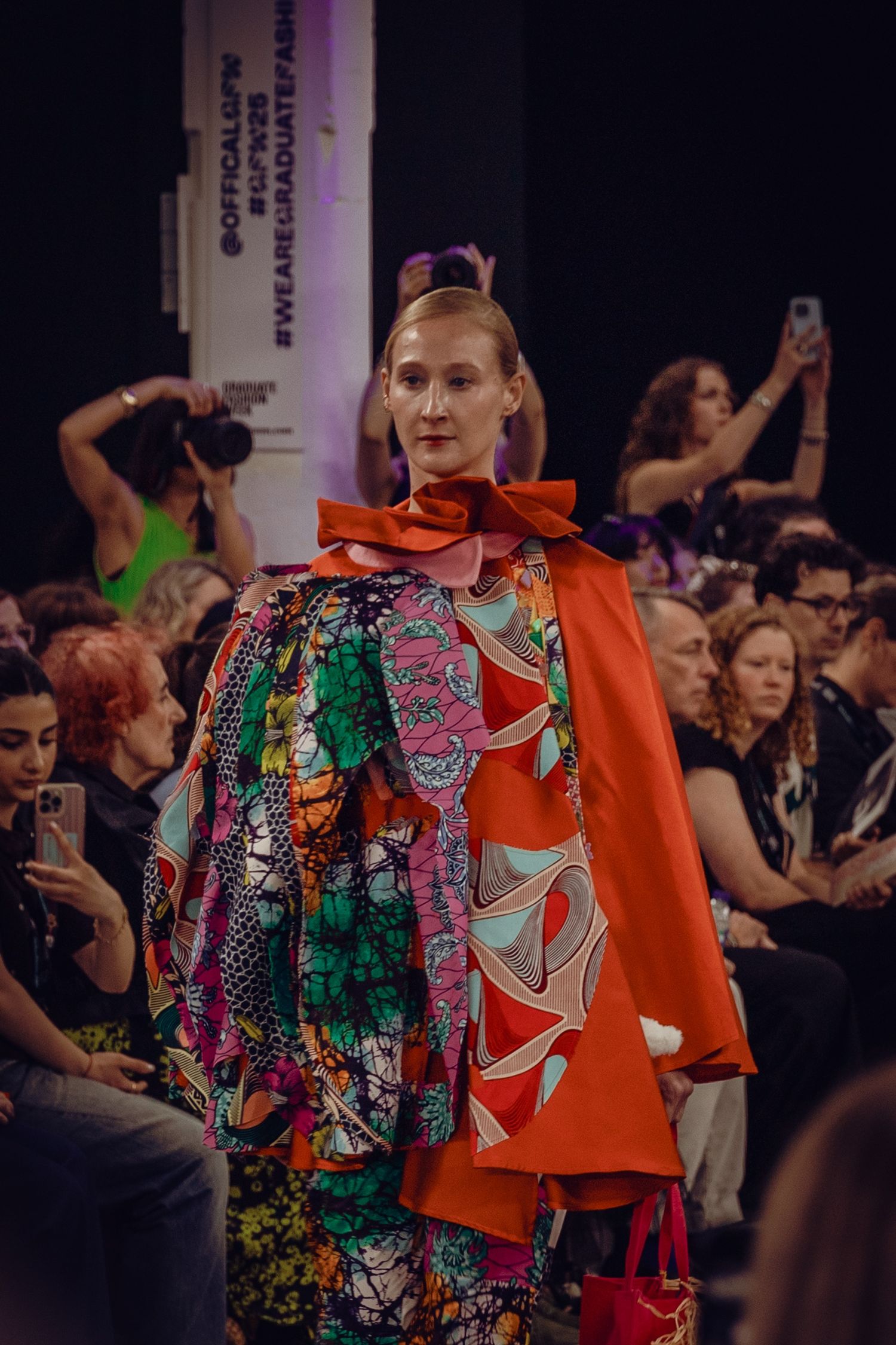 Fashion model walking runway in colorful patterned dress with dramatic orange cape at fashion show.