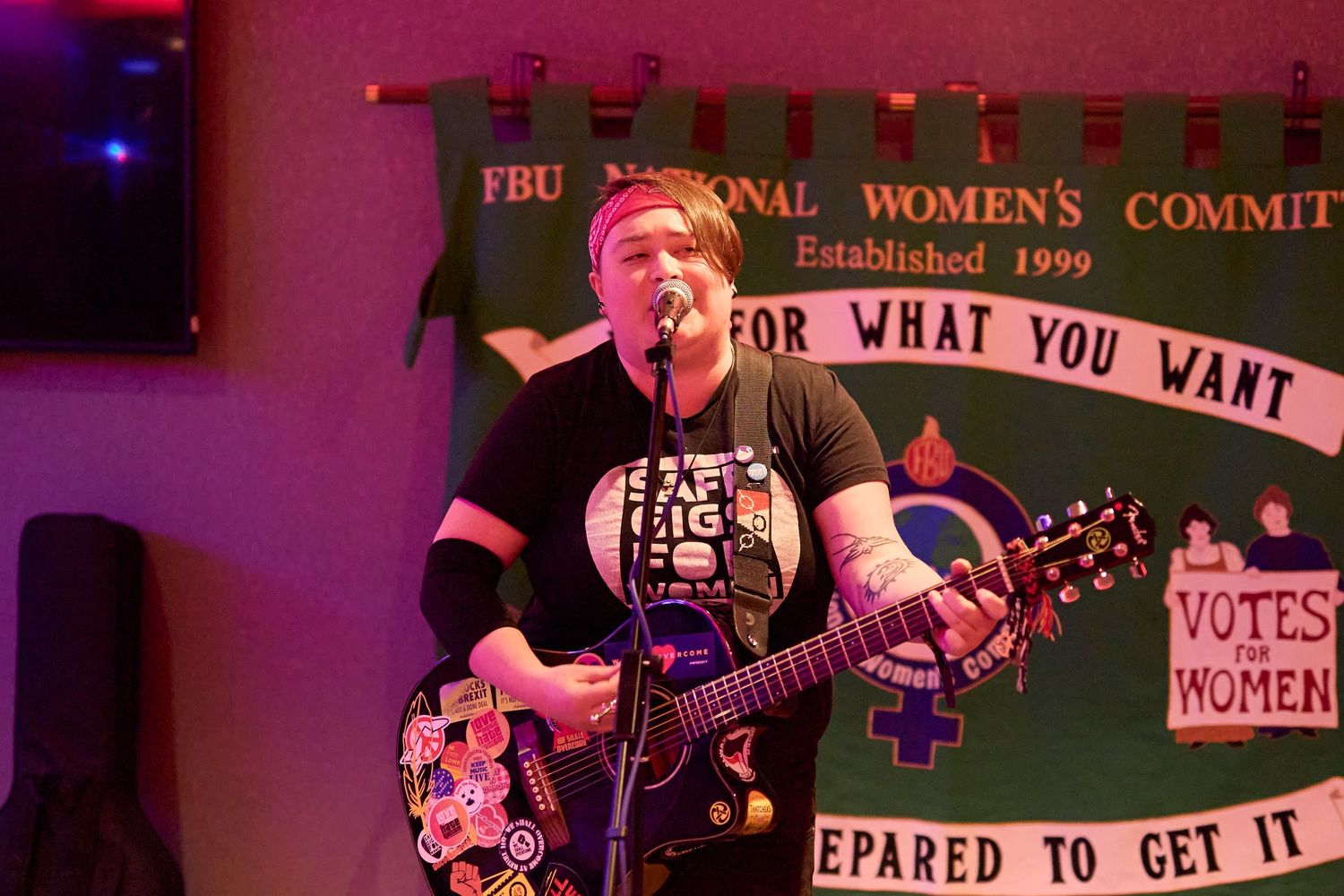 Musicians performing on stage with guitars at what appears to be a women's rights event with protest signs visible.