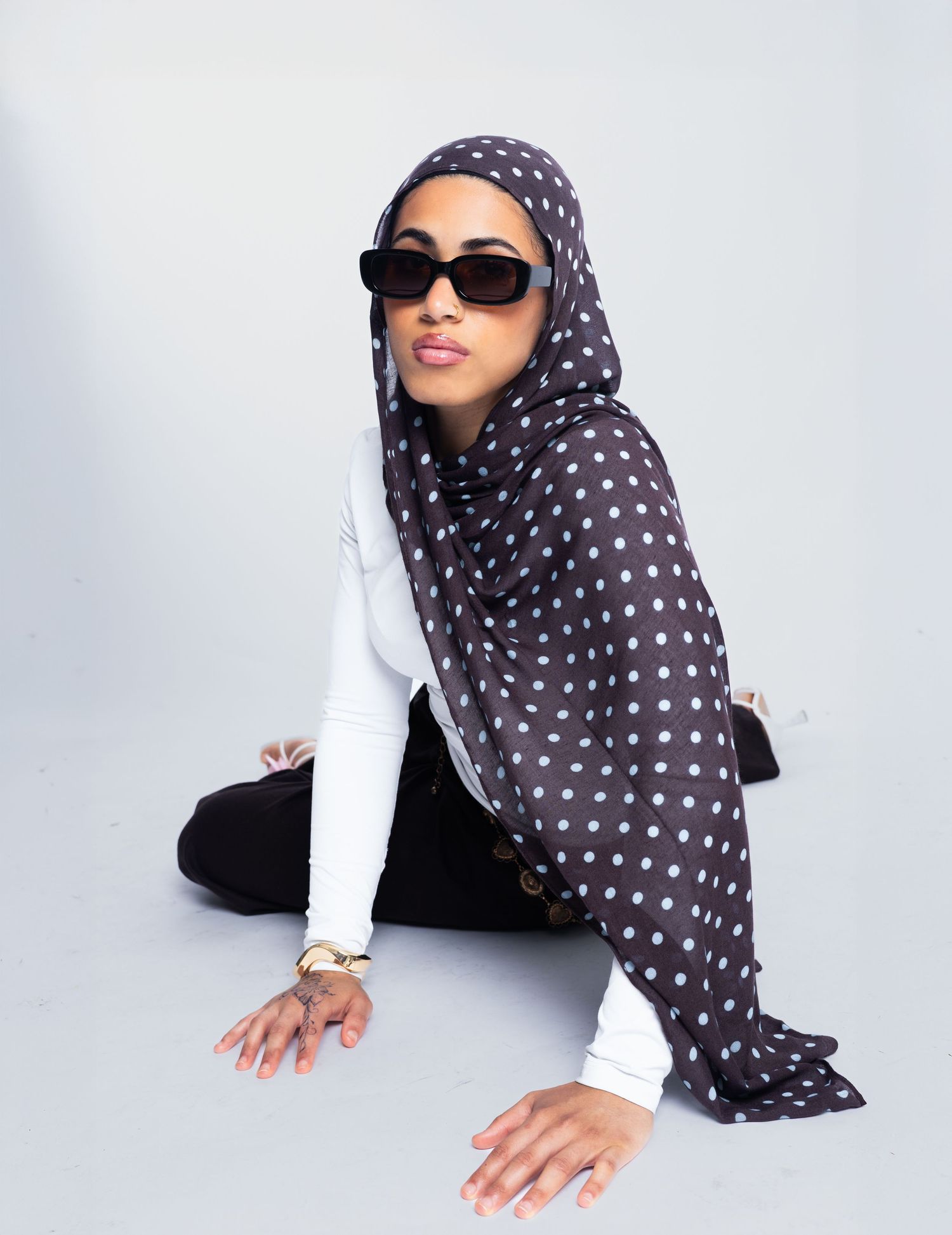 Woman wearing dark polka dot hijab, white top, black pants, and sunglasses, posing seated on white background.
