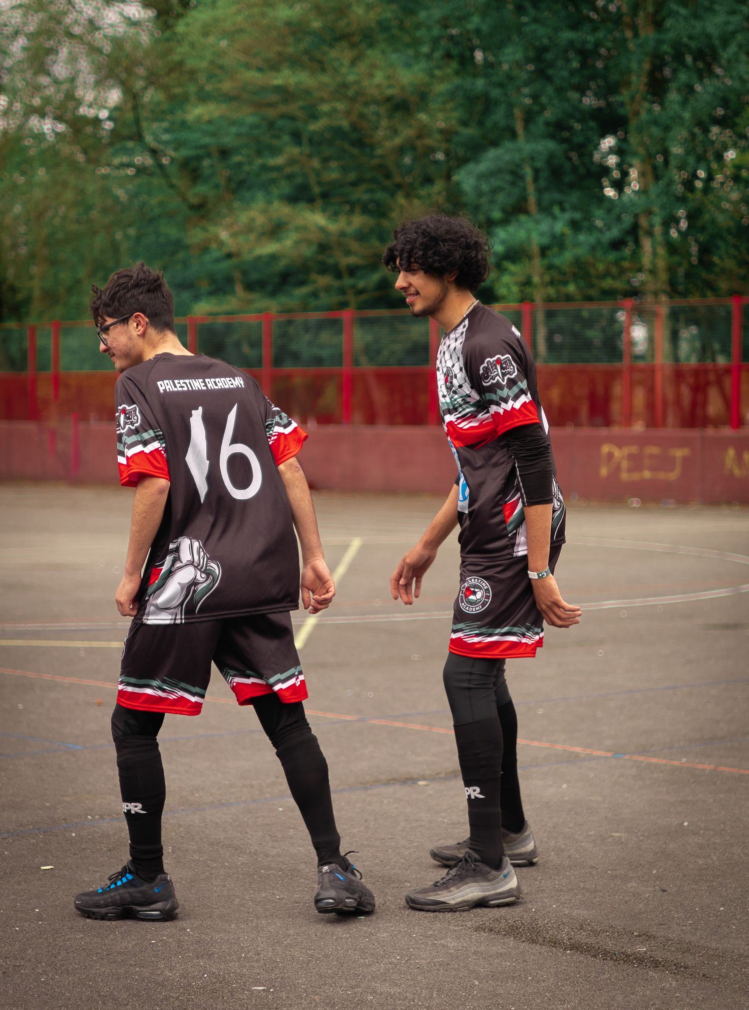 Soccer players in black and red uniforms standing on an outdoor court with trees in the background.