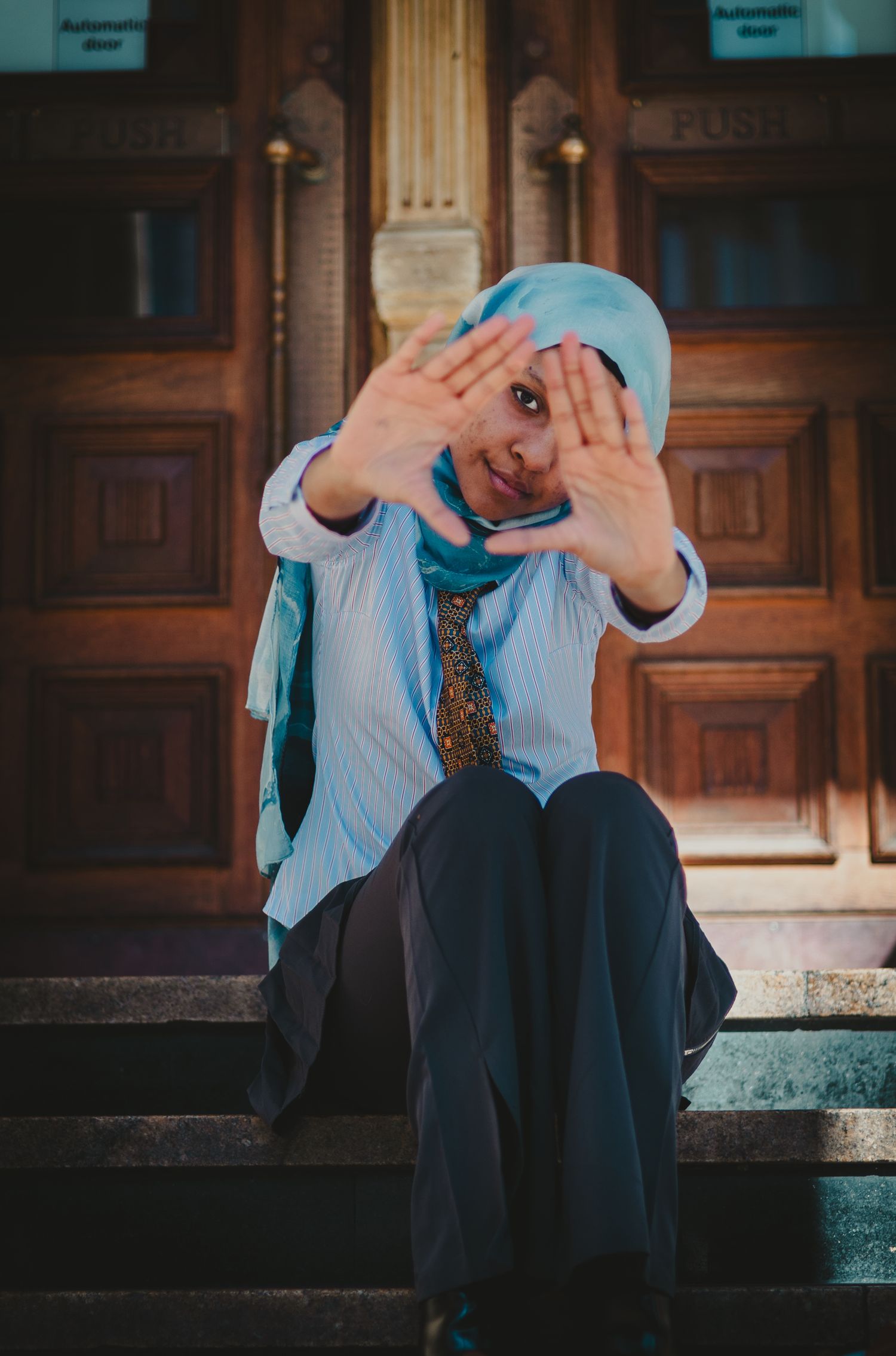 Person in business attire sitting on stone steps making playful hand gestures in front of wooden doors.