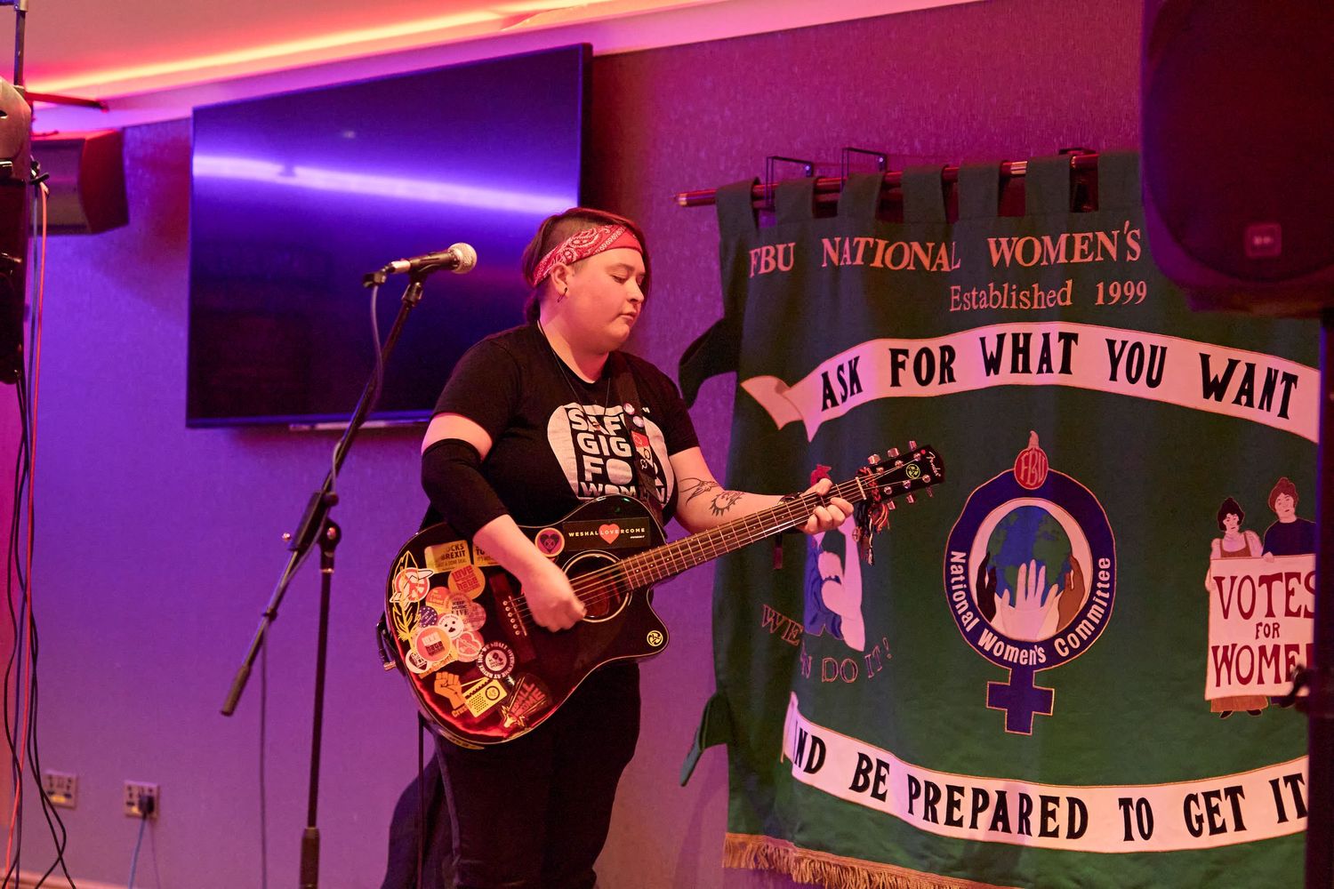 A musician performs with an electric guitar on stage with colorful purple and pink lighting and a banner in the background.
