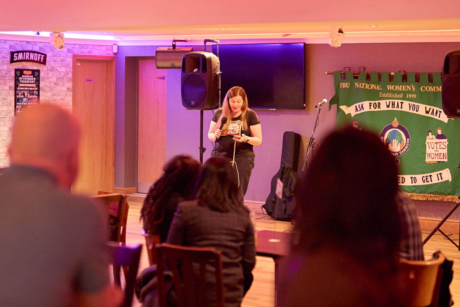 Person singing at a dimly lit karaoke venue with pink and purple mood lighting and TV screens displaying lyrics.