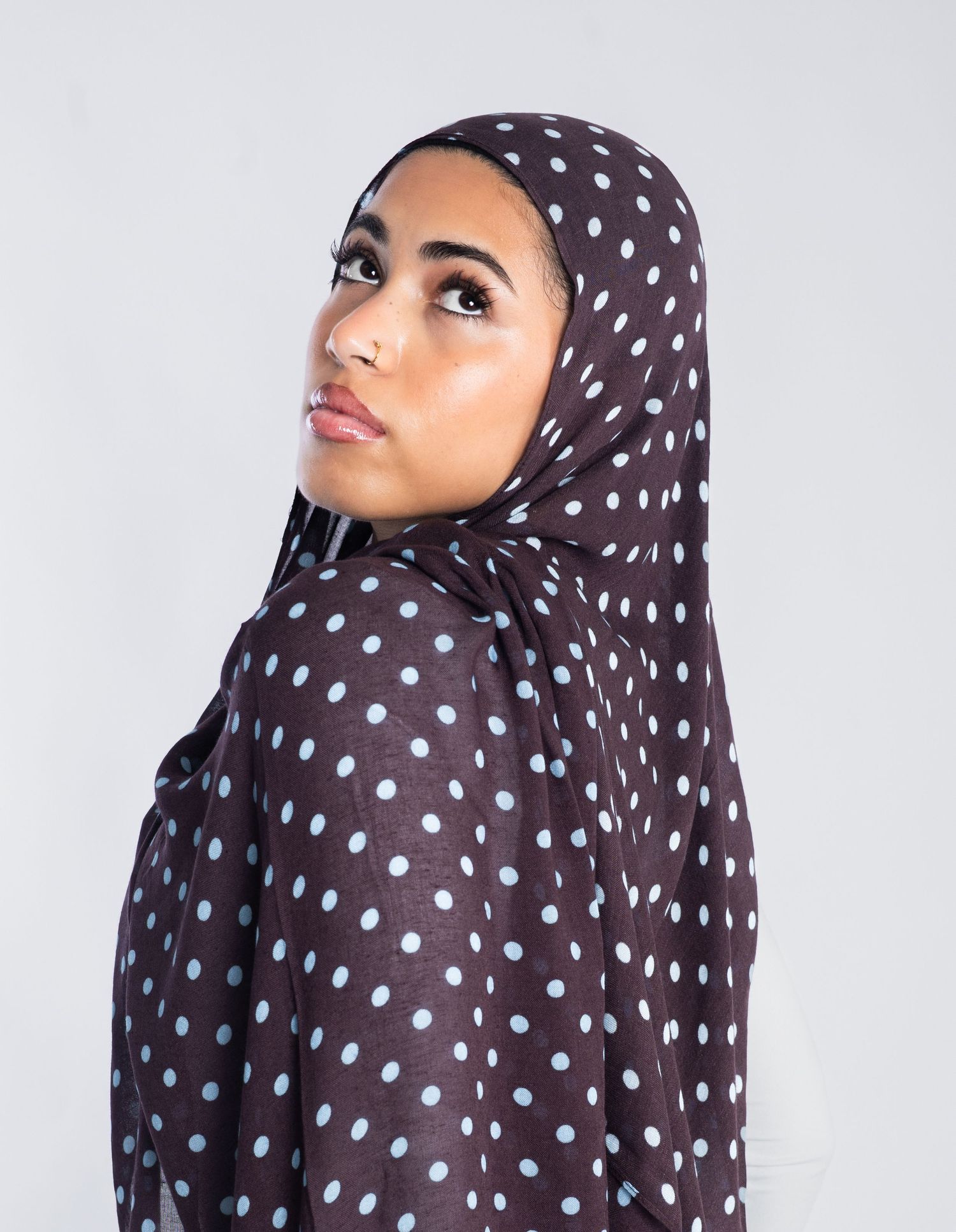 Woman wearing a dark brown polka dot hijab with light blue dots, posing against a light gray background.