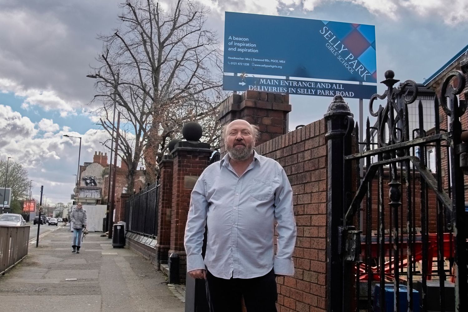 A person stands on a sidewalk next to a brick wall with a blue sign on a cloudy day.