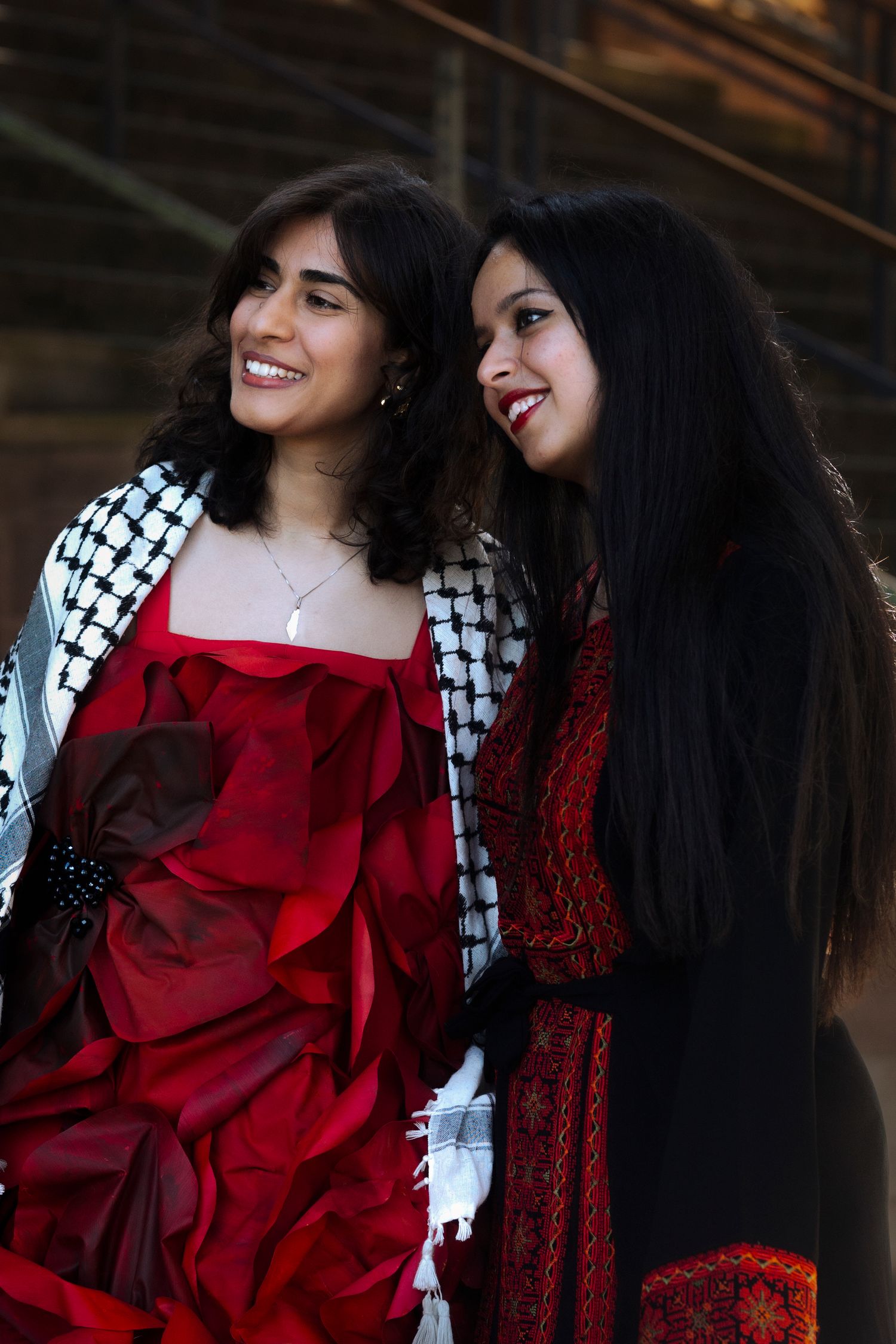 Two people wearing patterned scarves and red dresses pose together against stone building.