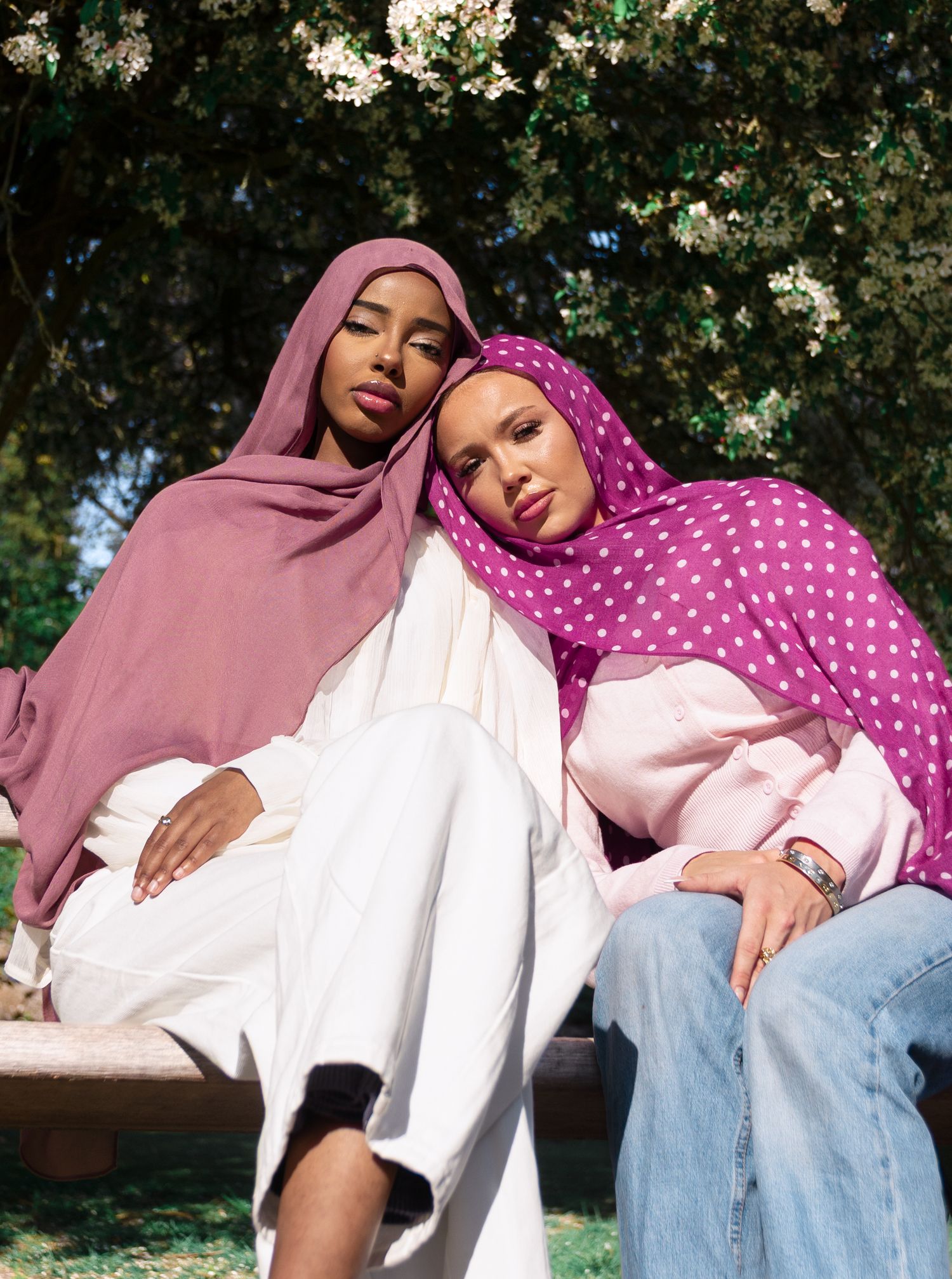 Two women wearing colorful hijabs sit outdoors under a flowering tree, one in dusty rose and one in polka dot magenta.