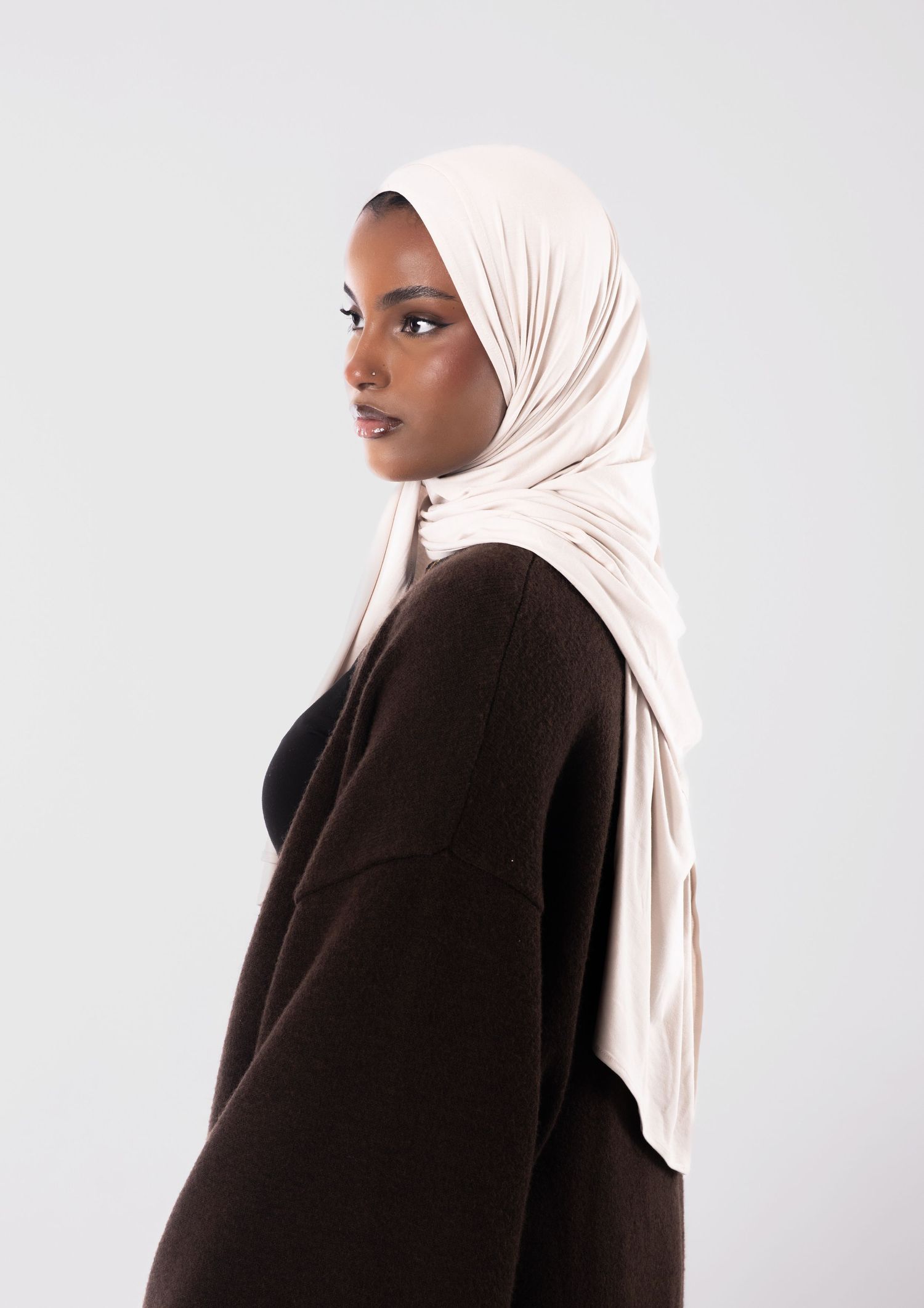 Woman wearing a cream hijab and dark brown knit cardigan, posed in profile against a light gray background.