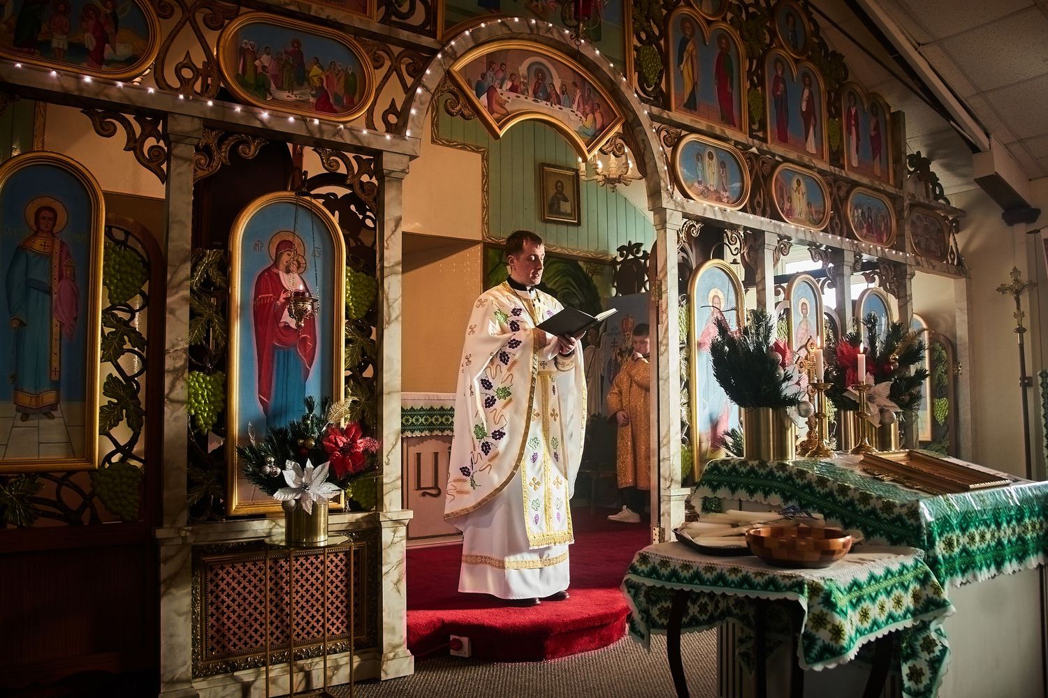 Ornate Eastern Orthodox church interior with iconostasis and priest conducting ceremony.