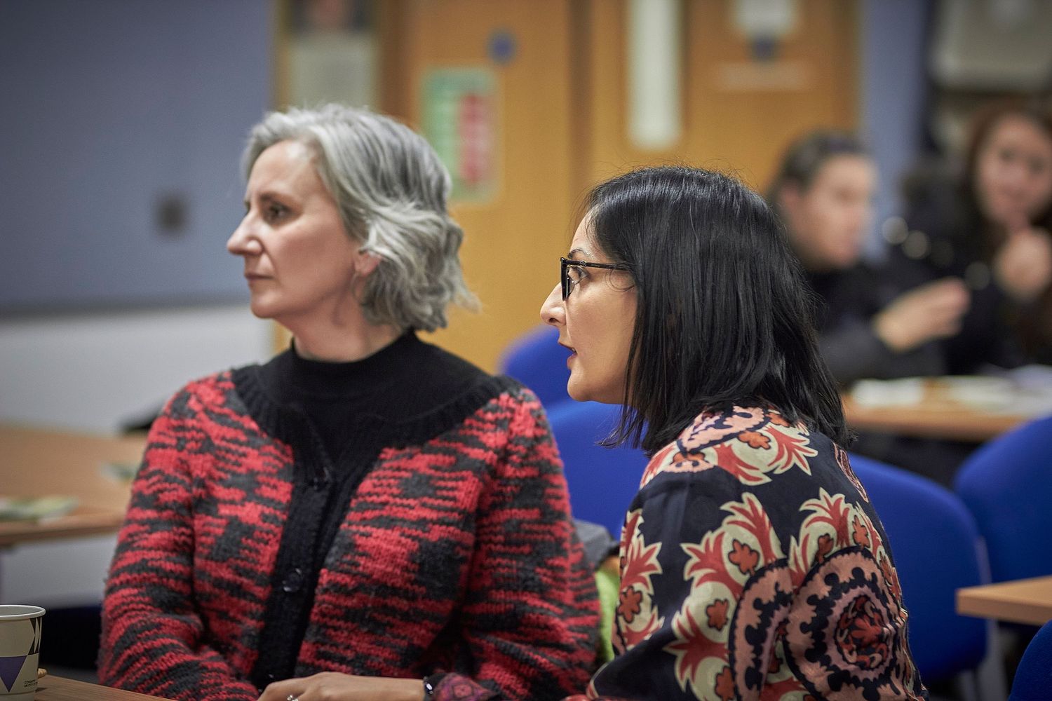 Two women sit together at a conference or lecture, wearing patterned red jackets in a classroom setting.