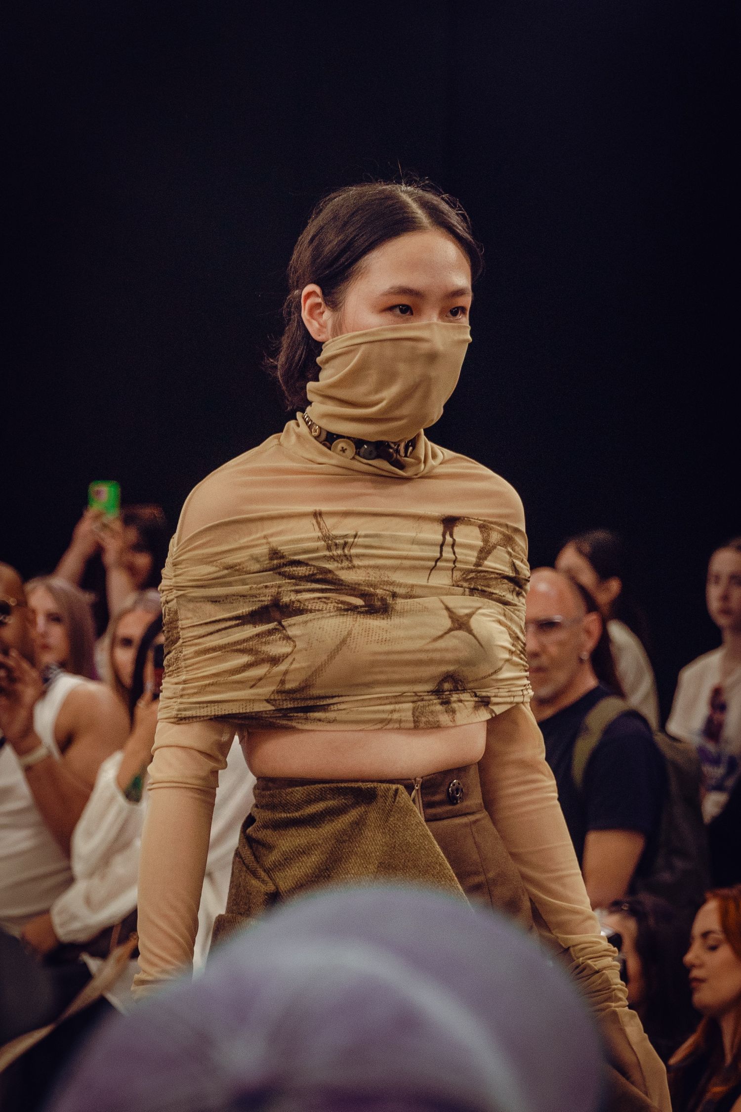 Model walks runway in beige wrapped crop top with face covering at fashion show with crowd in background.
