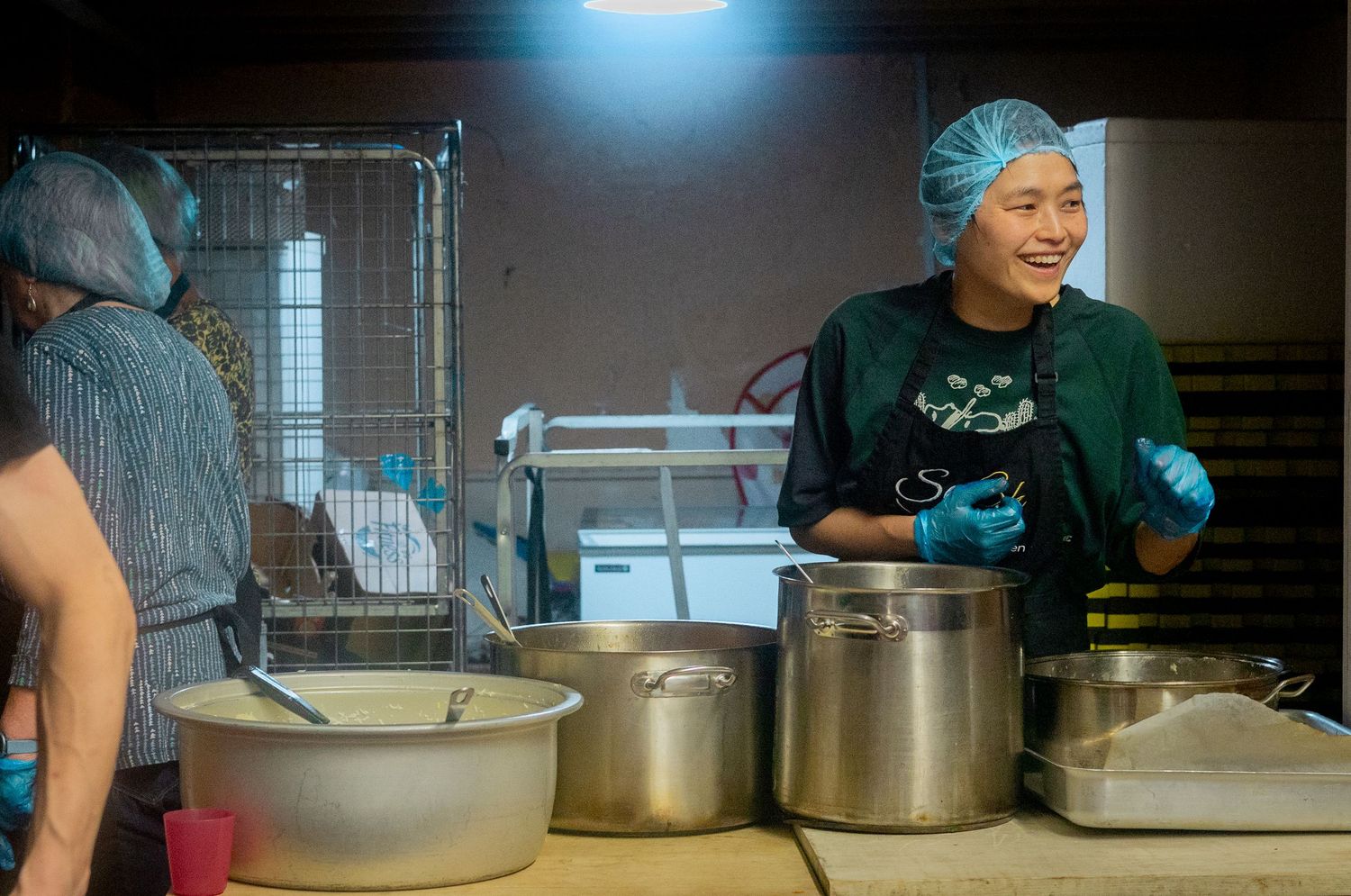 Volunteers in protective gear work together serving food from large silver pots.