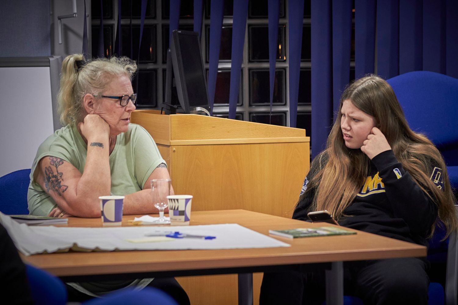 Two people having a discussion while sitting at a wooden table in what appears to be a classroom or meeting space.