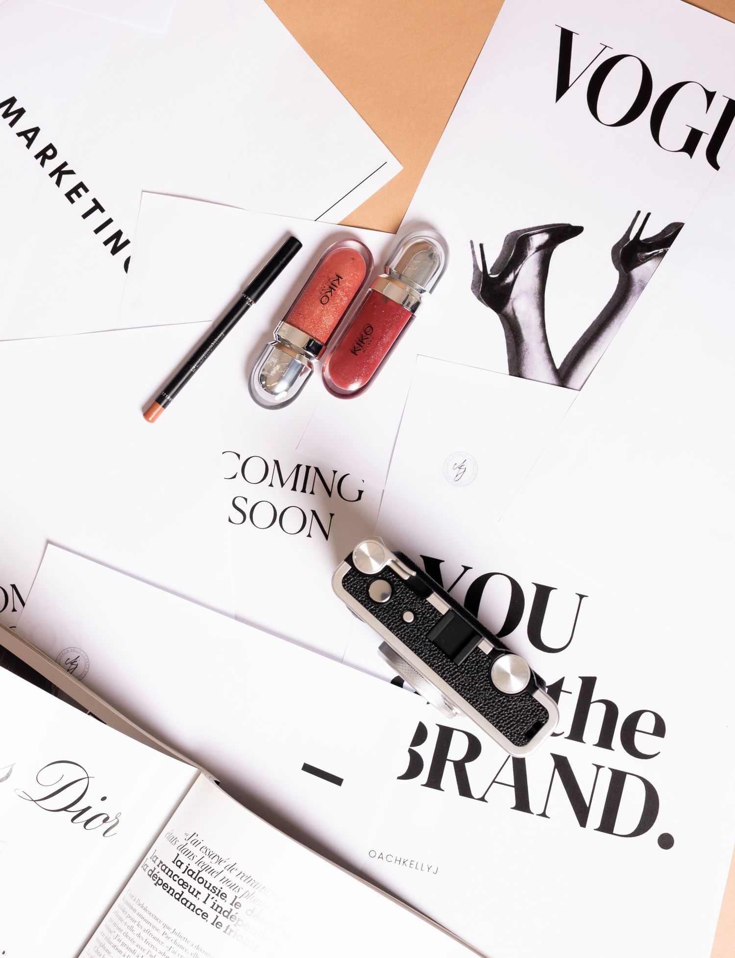 Flat lay of Vogue magazine, Dior cosmetics, lip liner, and brand marketing prints on white background.