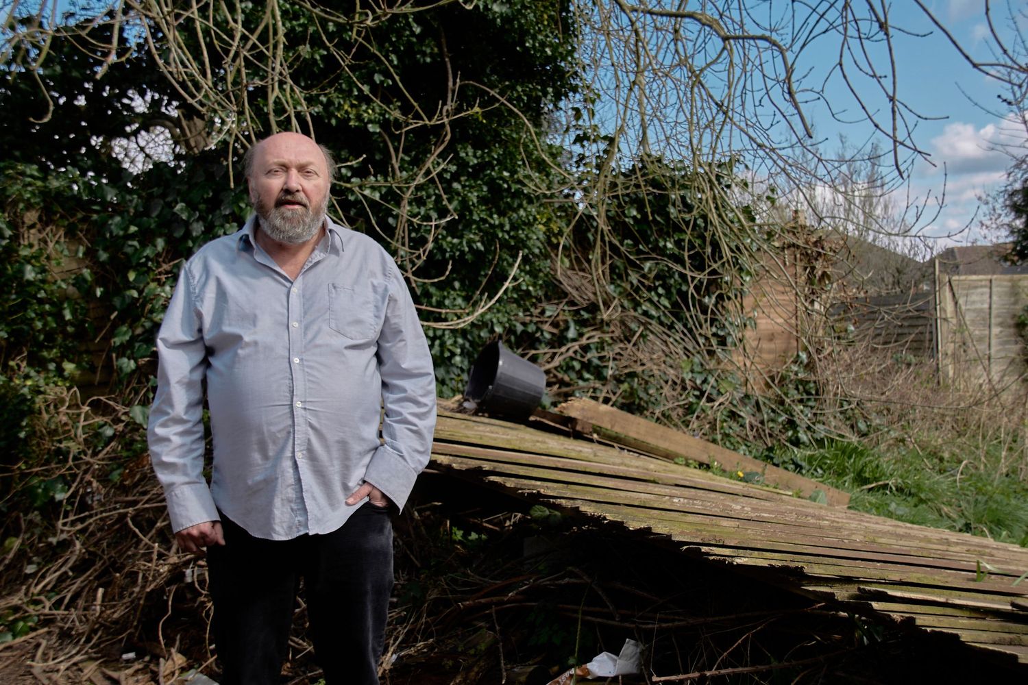 Person in gray shirt stands near pile of wooden planks against natural backdrop.