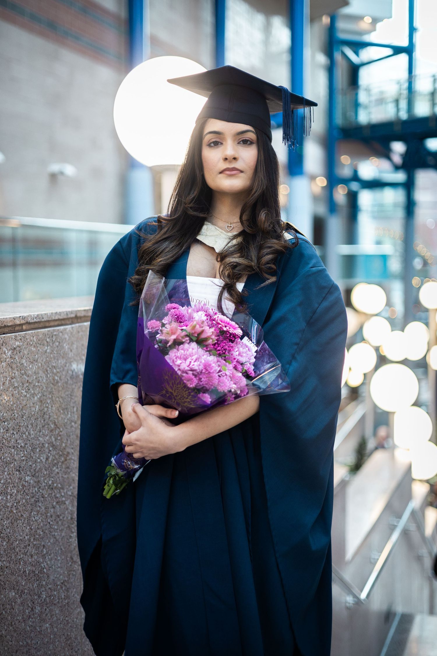 Graduate in dark blue cap and gown holds vibrant purple flower bouquet during graduation ceremony celebration.