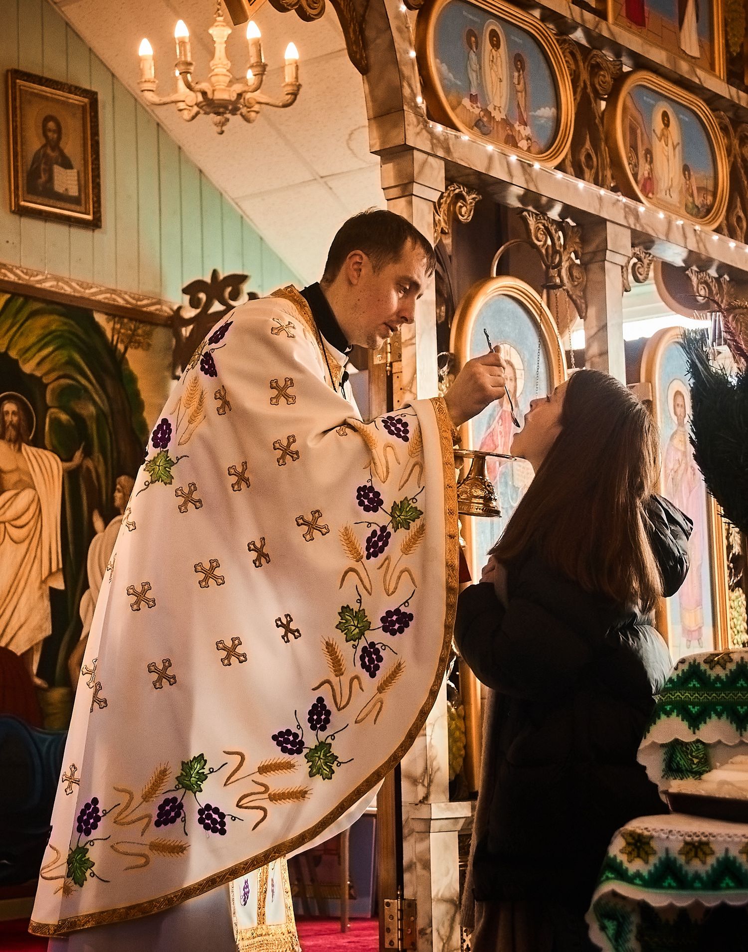 Orthodox priest in white vestments administering communion to a parishioner inside a decorated church.