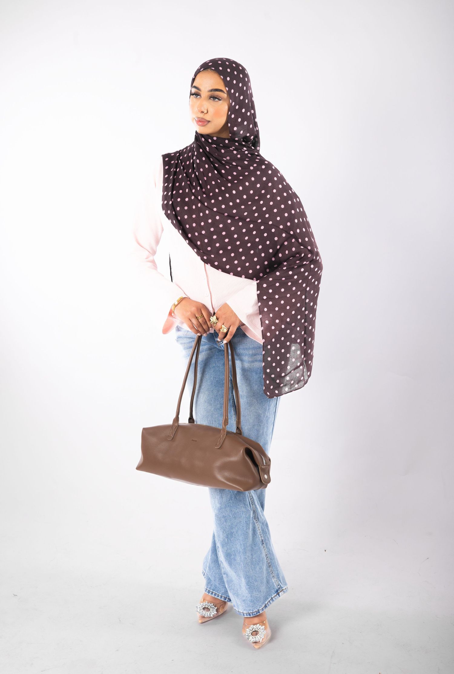 Woman wearing brown polka dot hijab, pink top, jeans, holding brown handbag, standing against white background.