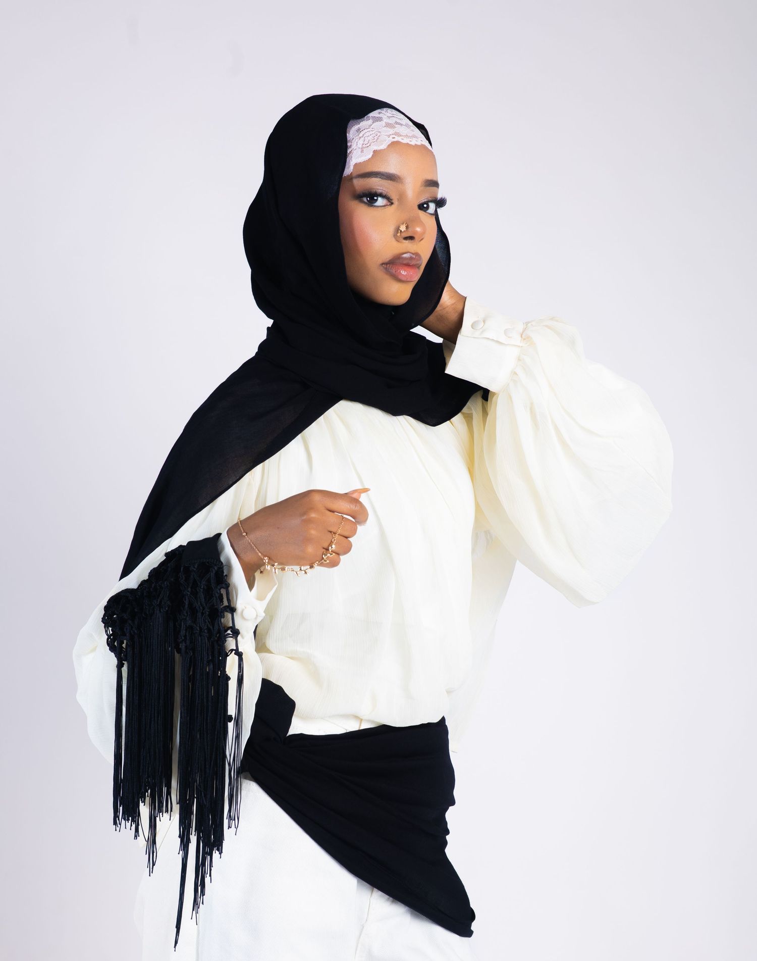 Woman wearing a black hijab with fringe and white puff-sleeve top poses gracefully against a light background.