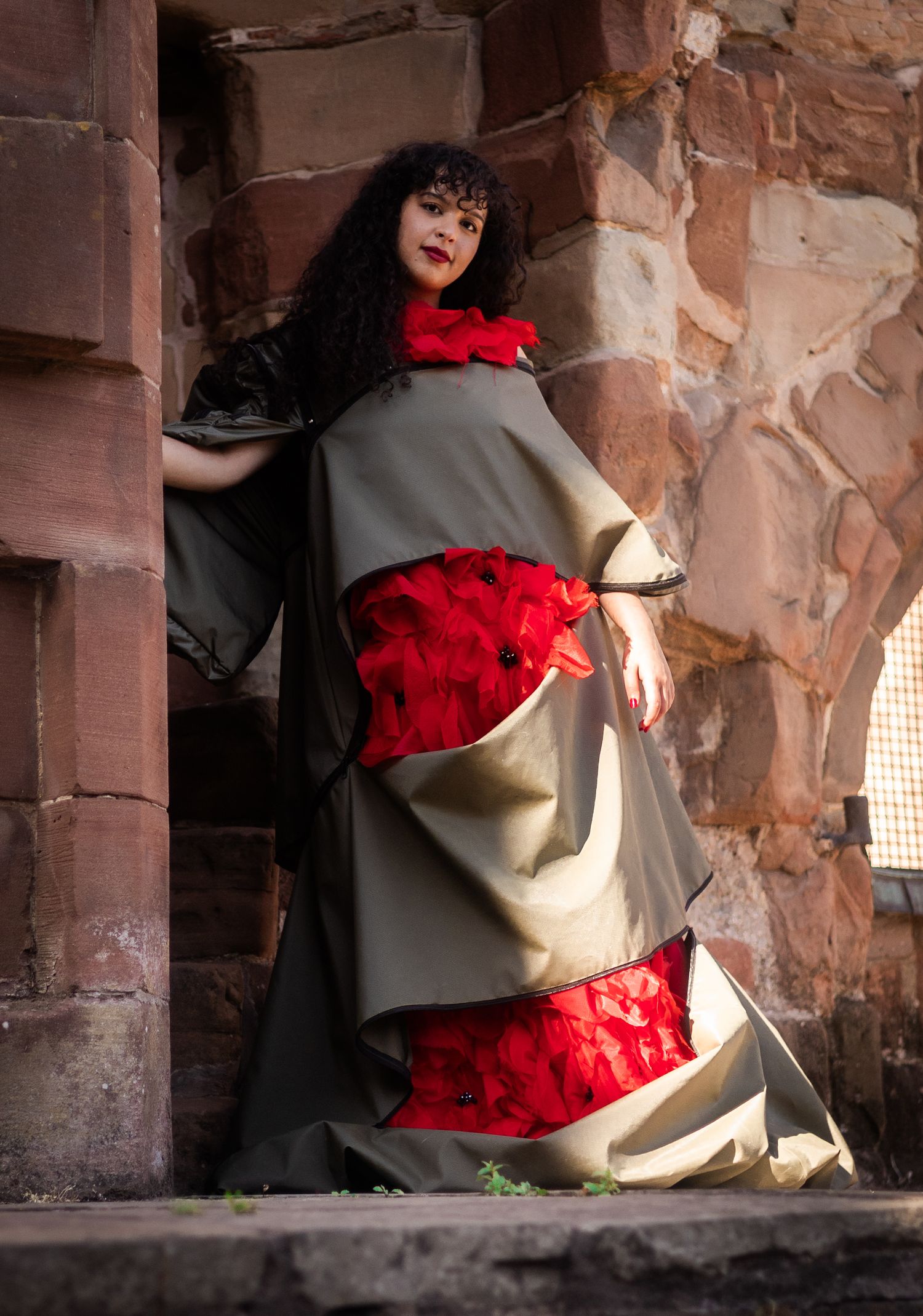 Dramatic fashion portrait showing flowing grey cape and red ruffled dress against stone wall background.