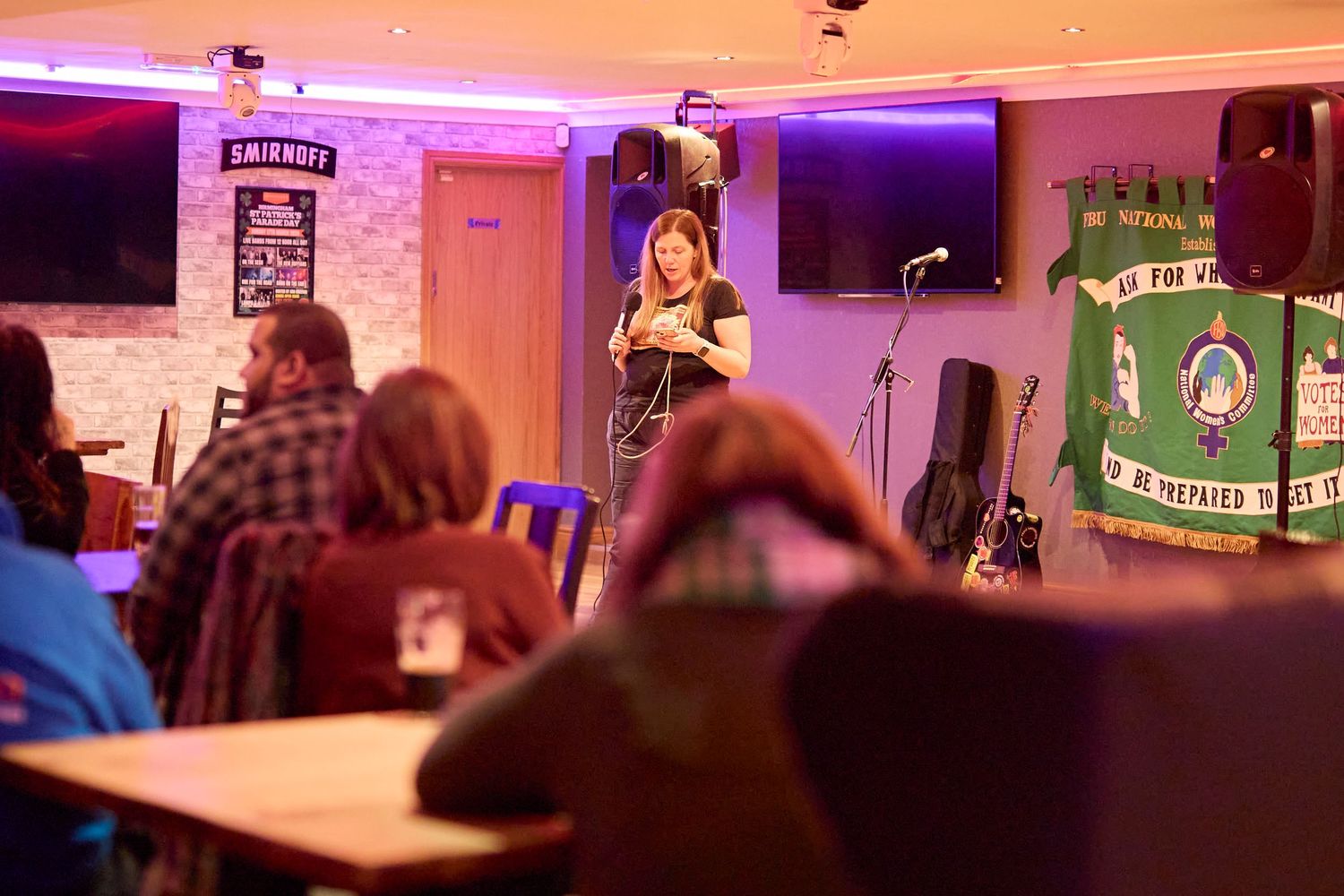 A performer stands on stage at a small music venue bathed in purple lighting with audience members seated in the foreground.