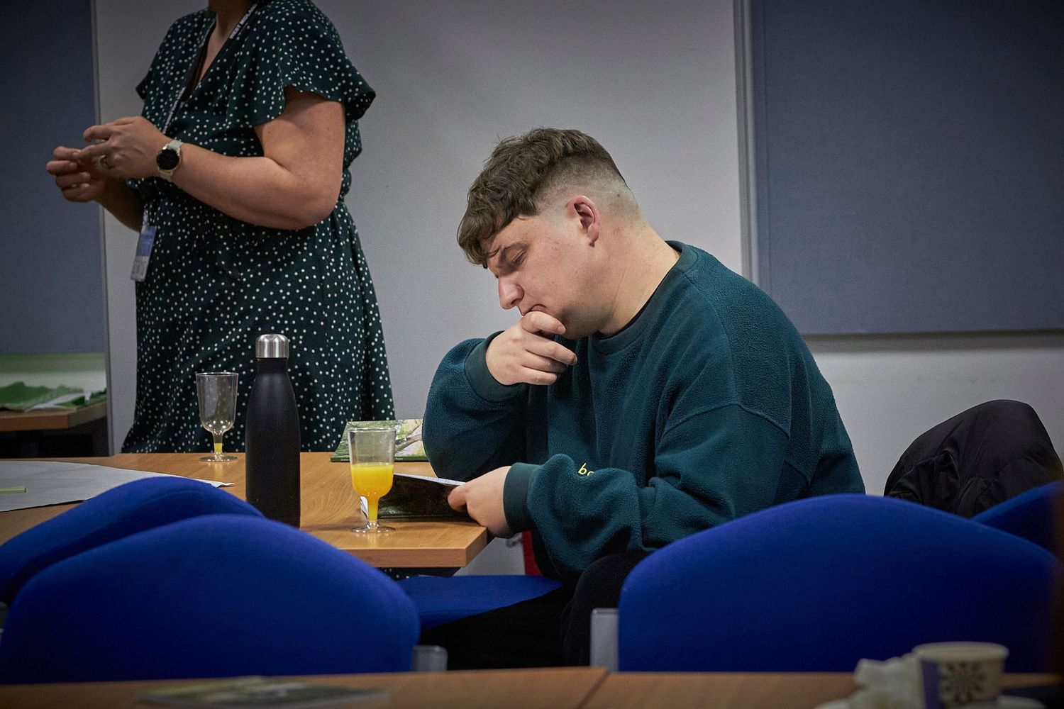 Student wearing dark green sweatshirt sits thoughtfully at desk in classroom with blue chairs while someone stands nearby.