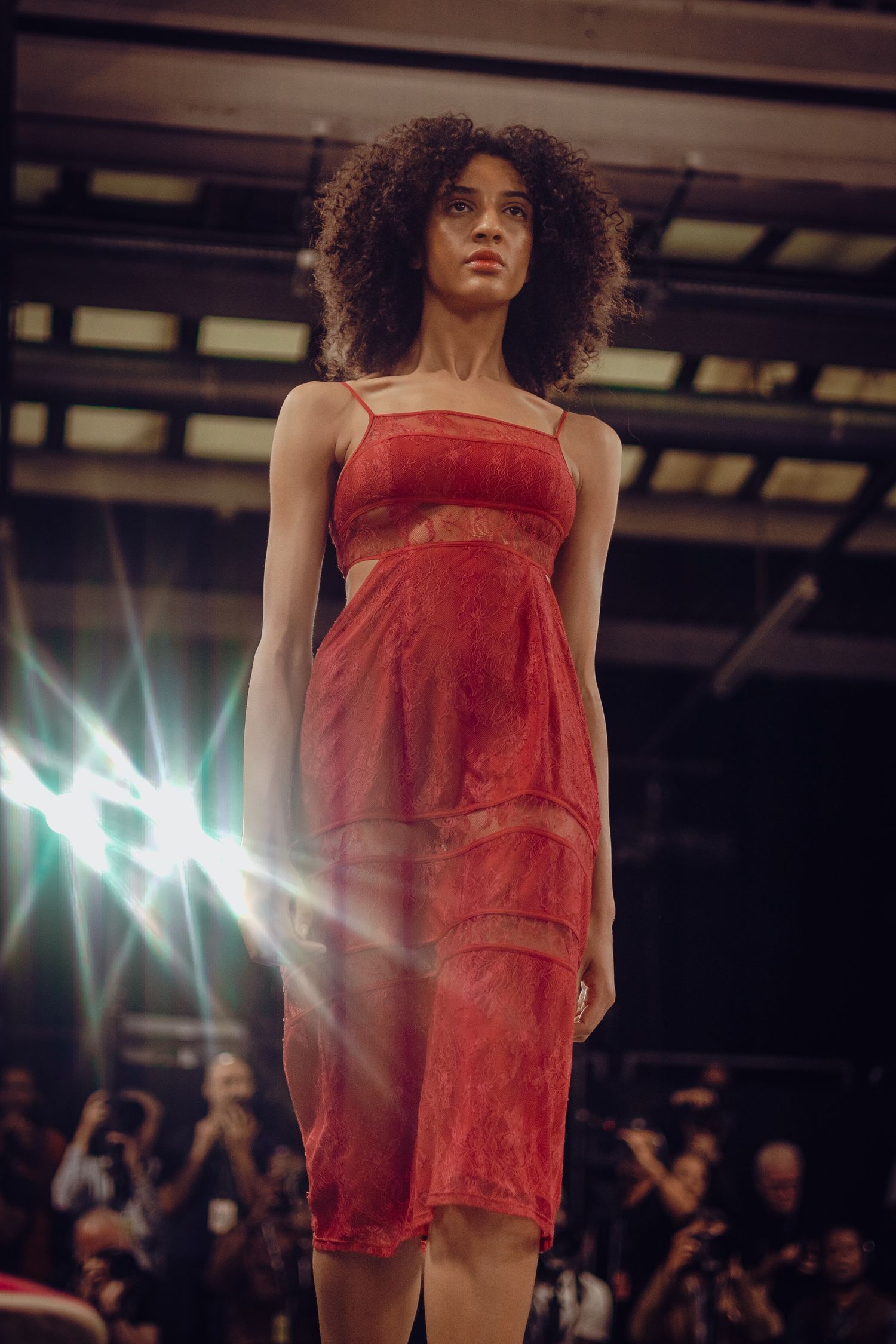 Model in red fitted dress walking runway with dramatic spotlight effects in background.