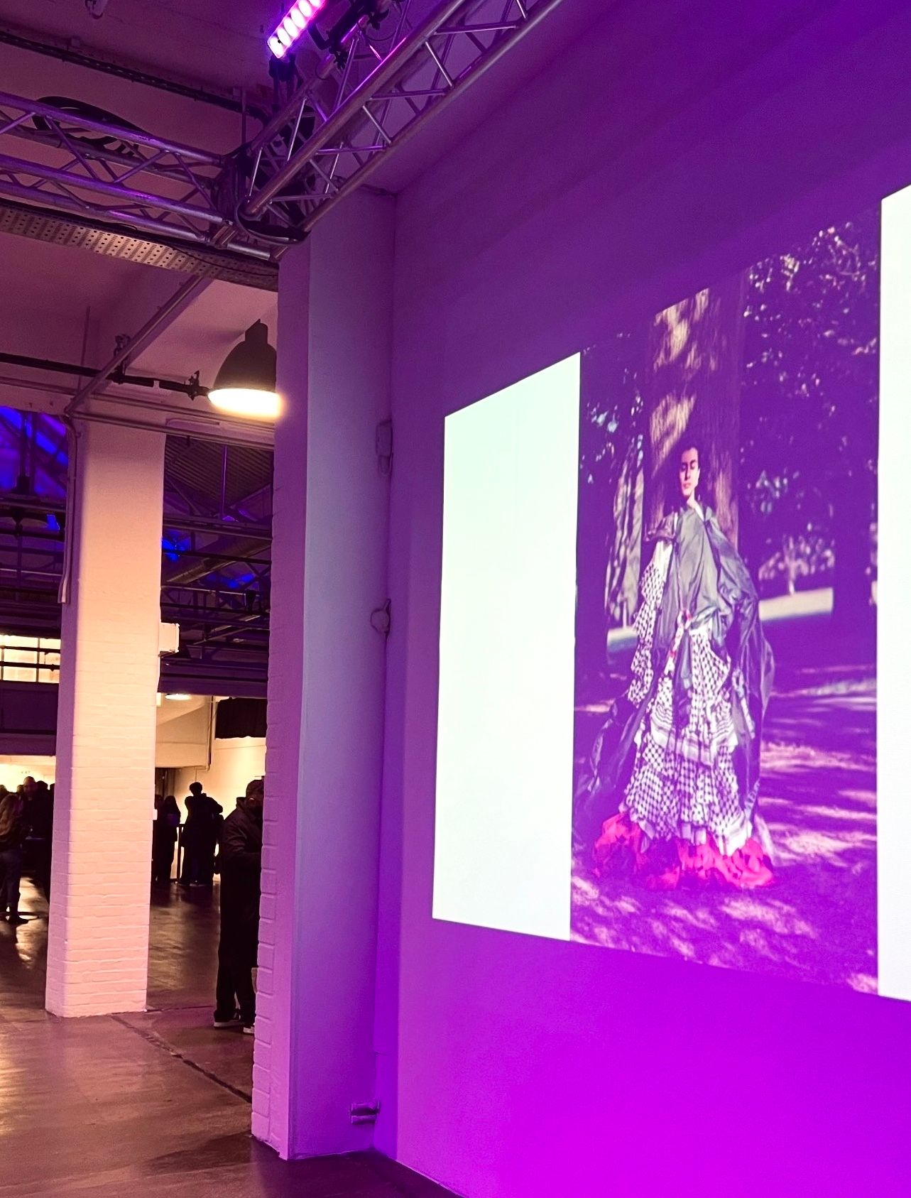 Purple-lit gallery space shows projected images on white walls with industrial ceiling visible above.