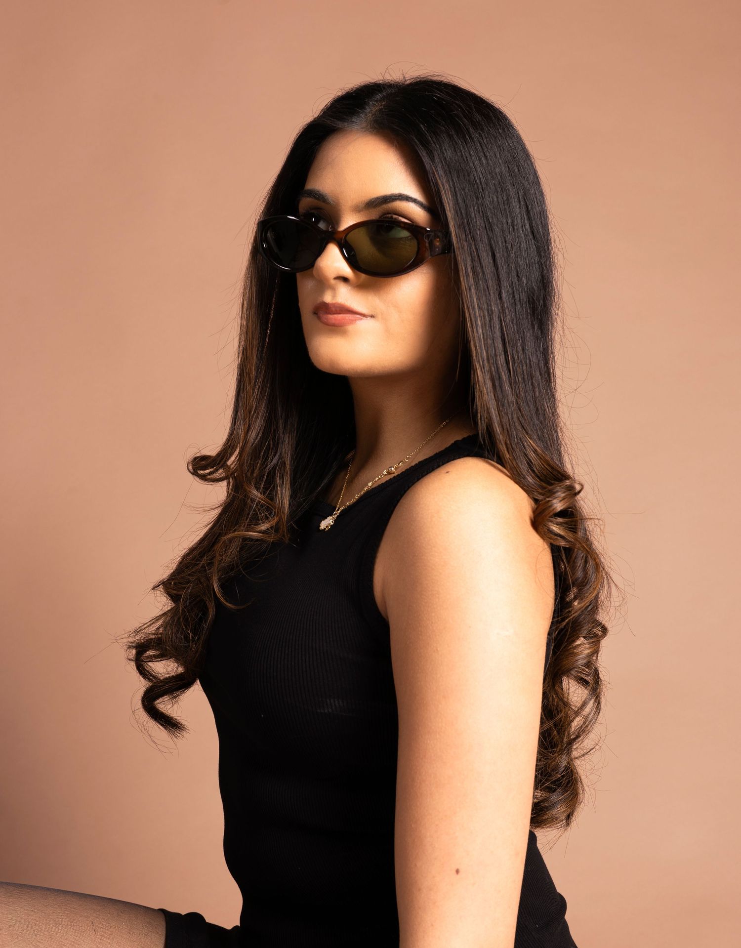 Young woman with long dark hair wearing black sunglasses and a black sleeveless top, posing against a beige background.