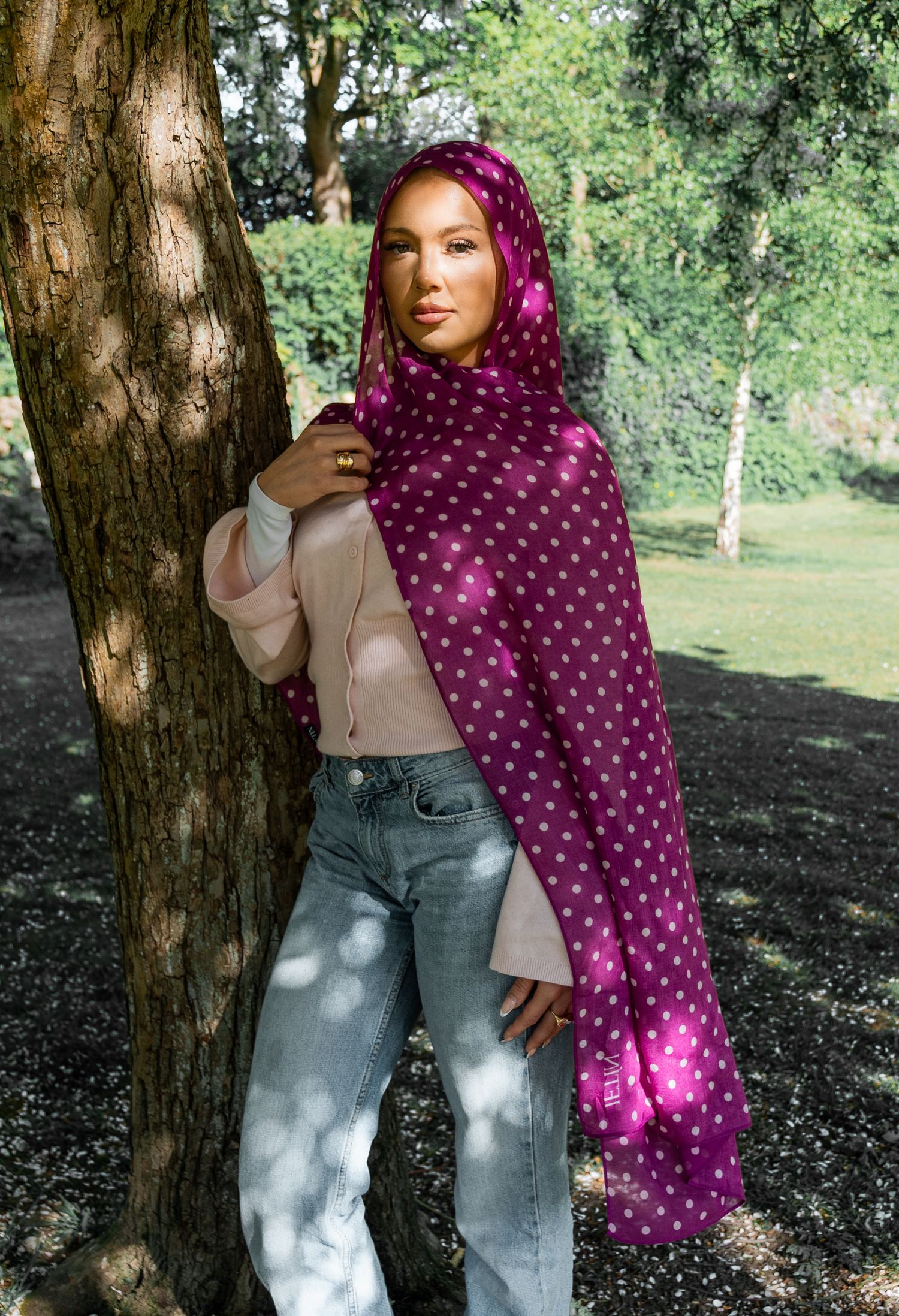 Woman wearing a magenta polka dot hijab and scarf with jeans, leaning against a tree in a park.