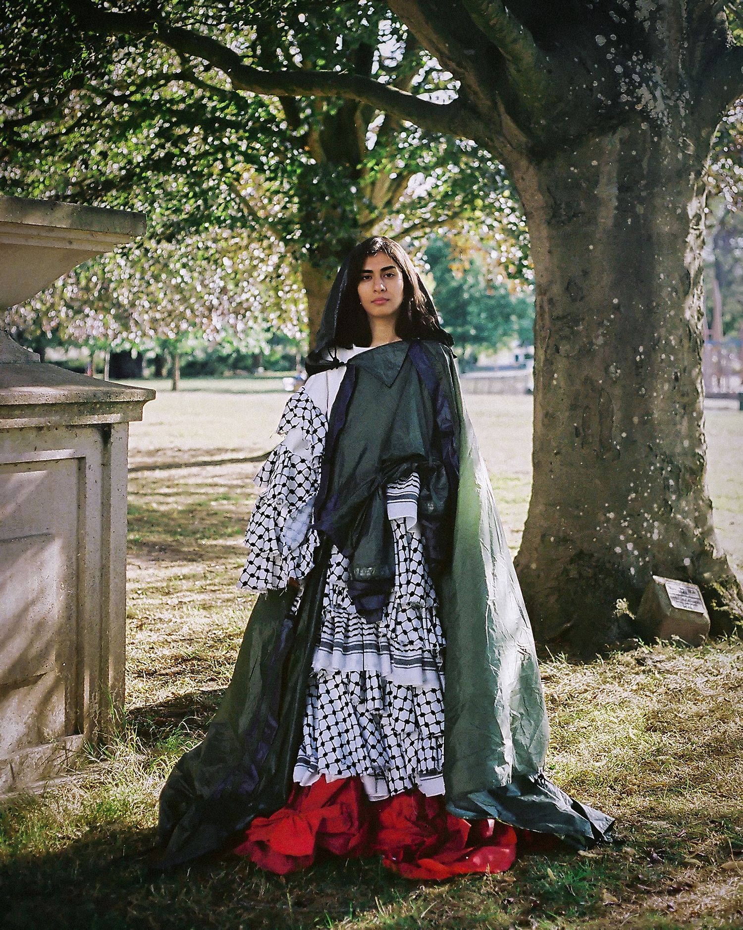 Woman in layered keffiyeh-patterned gown with green and red fabric stands under a tree in a park.