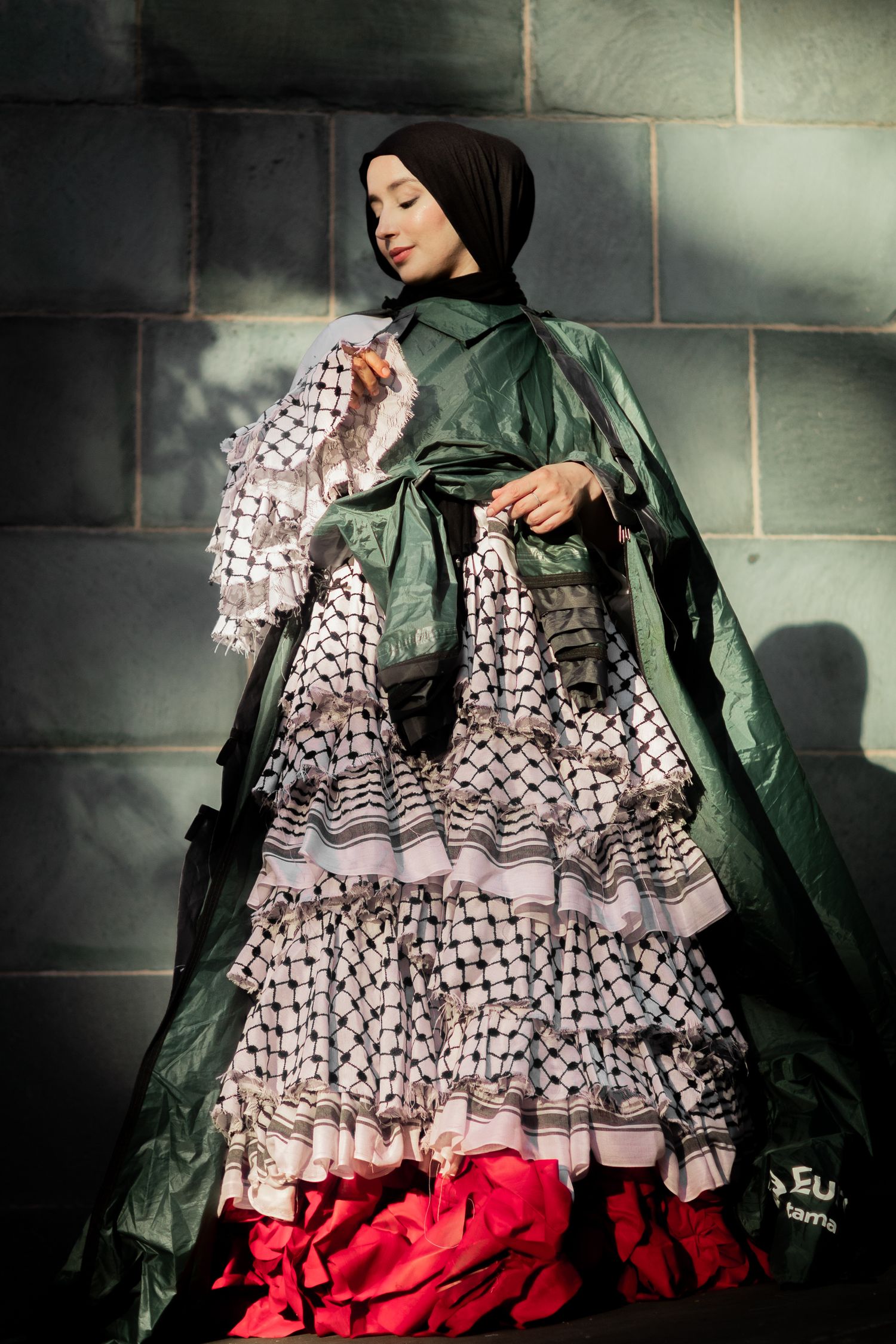 Dramatic fashion portrait showing flowing green cape and black and white ruffled dress with red accents against stone wall.