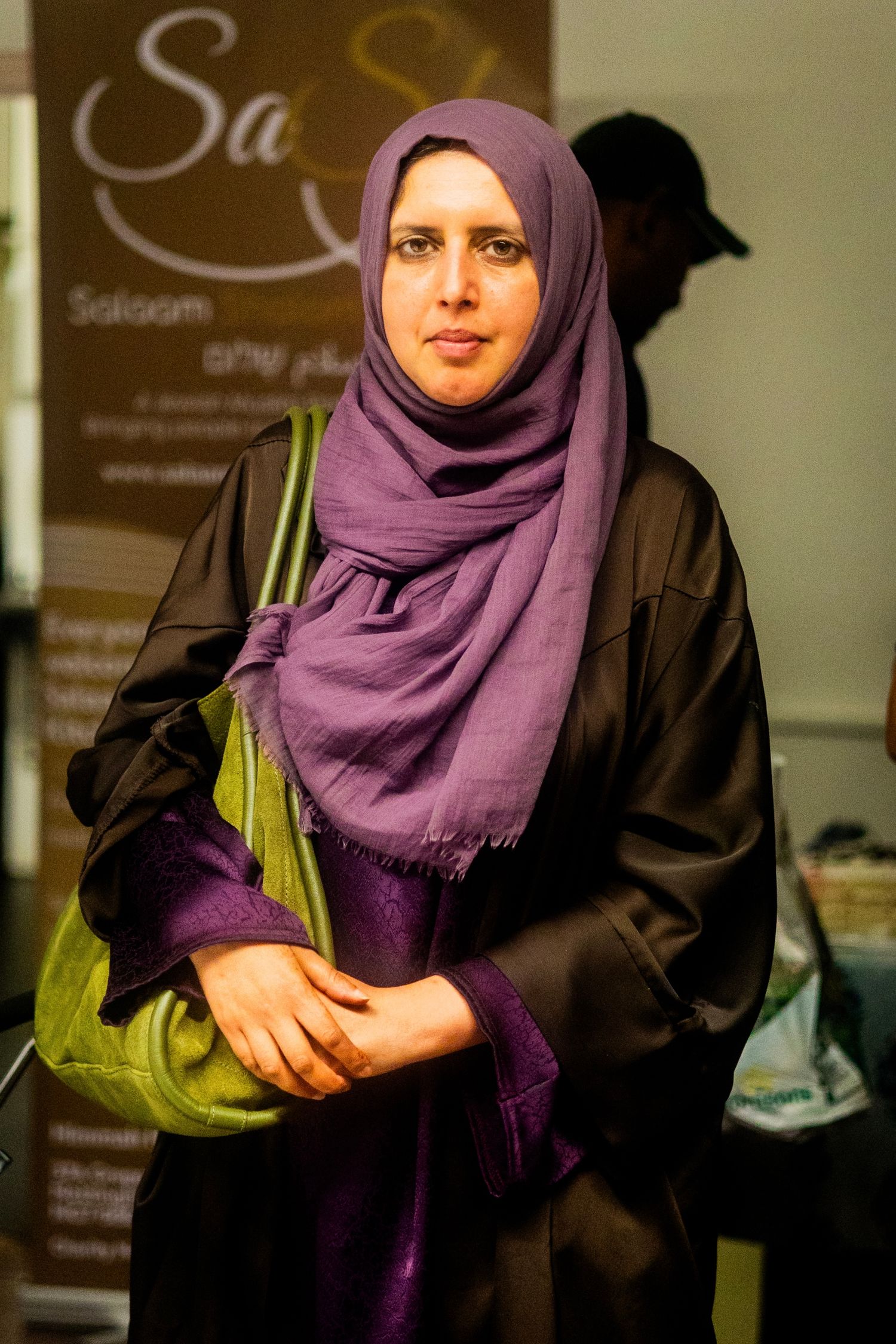 Figure in purple hijab and dark clothing stands in indoor setting with signage.