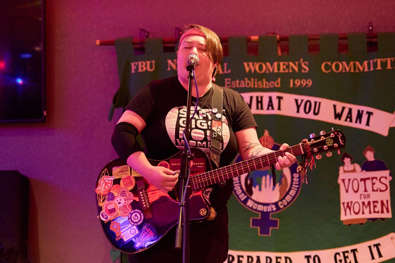 A performer plays guitar and sings on stage with a feminist banner visible in the background.