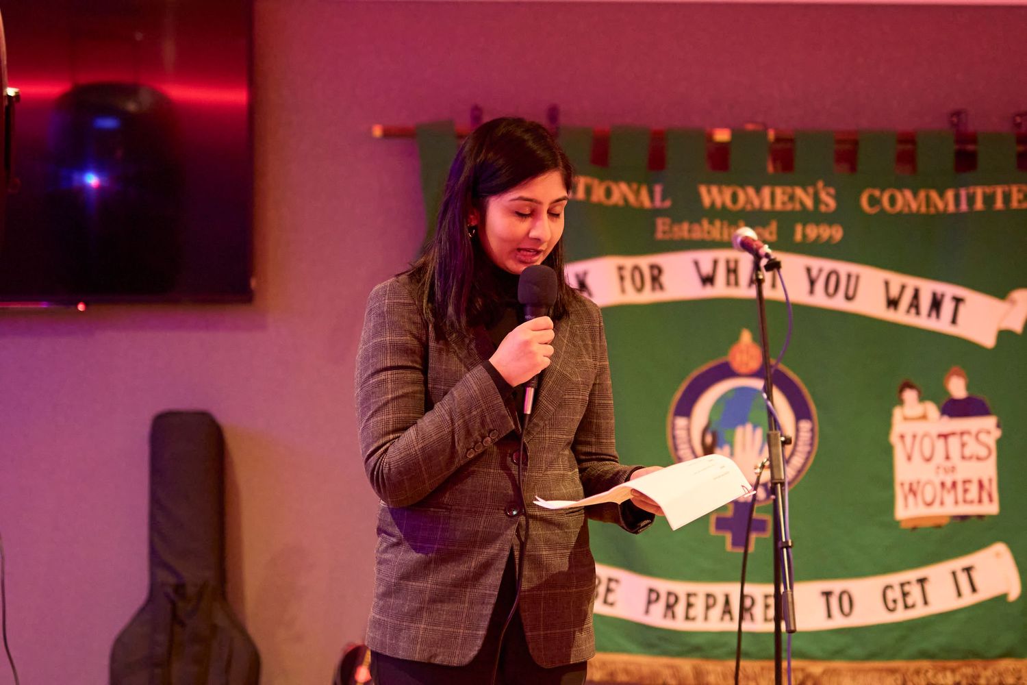 A person in a brown jacket speaks into a microphone at a podium with a green banner in the background.