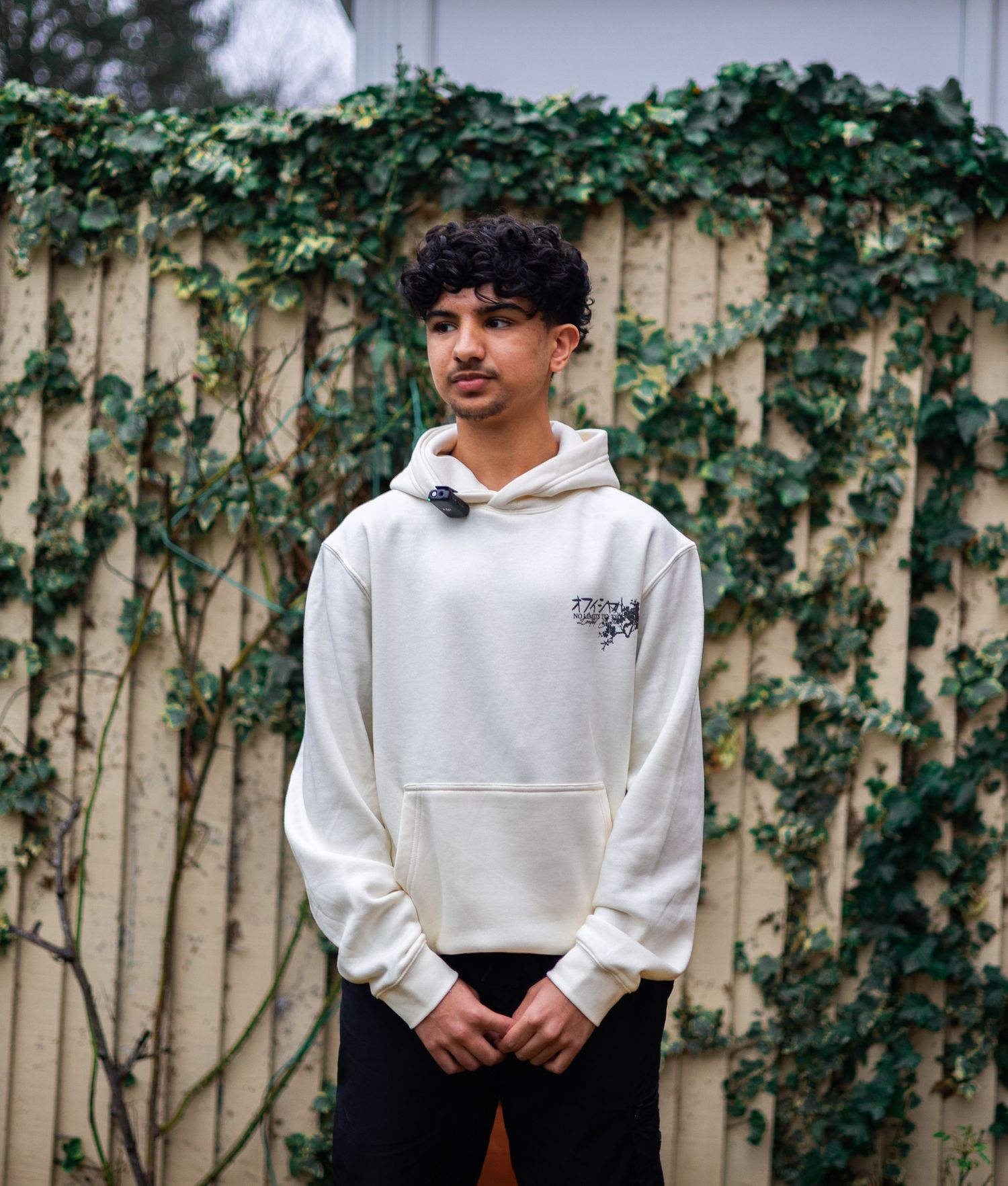 Young man in white hoodie standing before ivy-covered wooden fence outdoors.