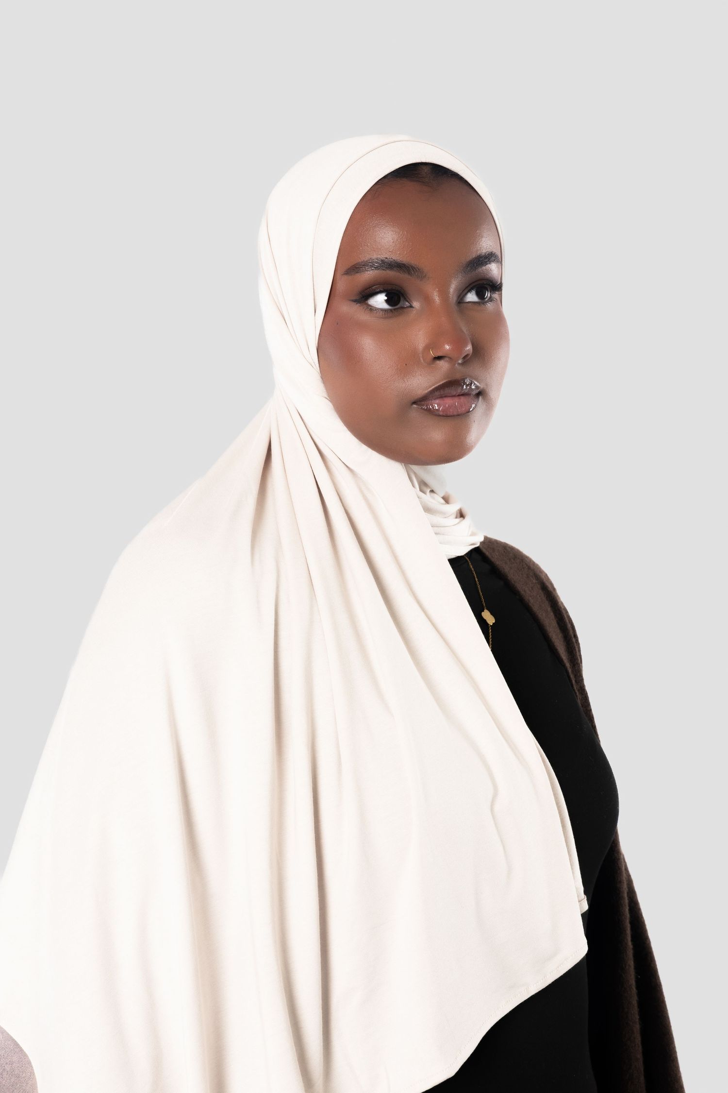 Young Black woman wearing a cream hijab and black outfit, posing against a light gray background.