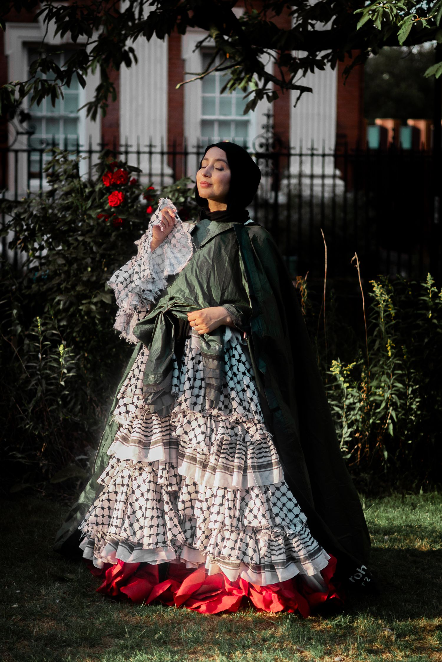 A flowy polka dot dress with red trim and green cape is worn in a garden setting with architectural details.