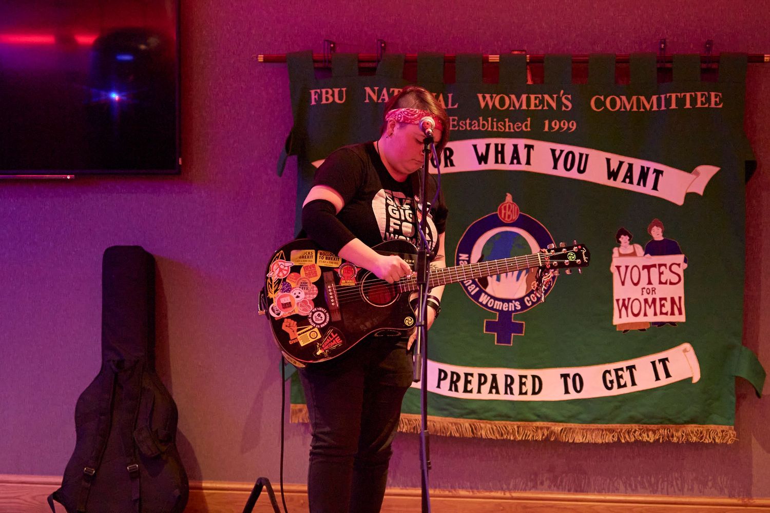 A performer stands with a guitar on stage with a pink-lit backdrop featuring feminist protest banners and slogans.
