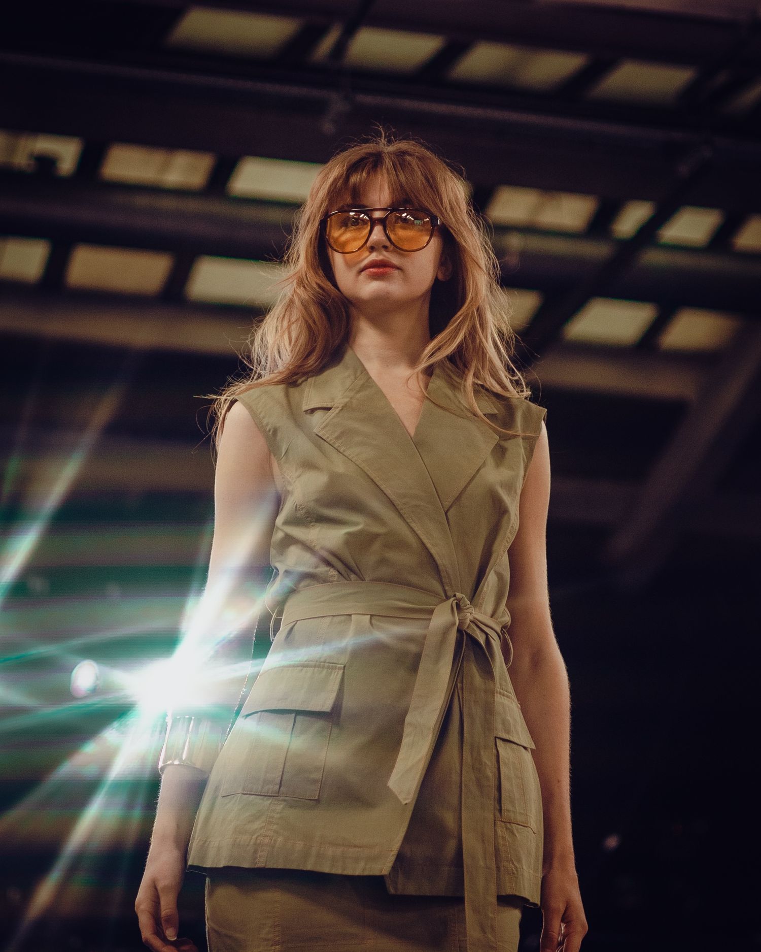 Model wearing beige sleeveless belted blazer vest with glasses on fashion runway under dramatic lighting.