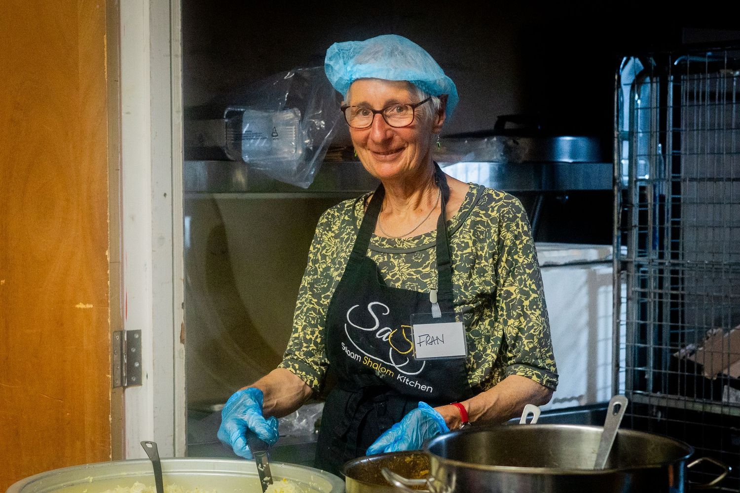 A volunteer in protective gear serves food from large pots in an industrial kitchen setting.