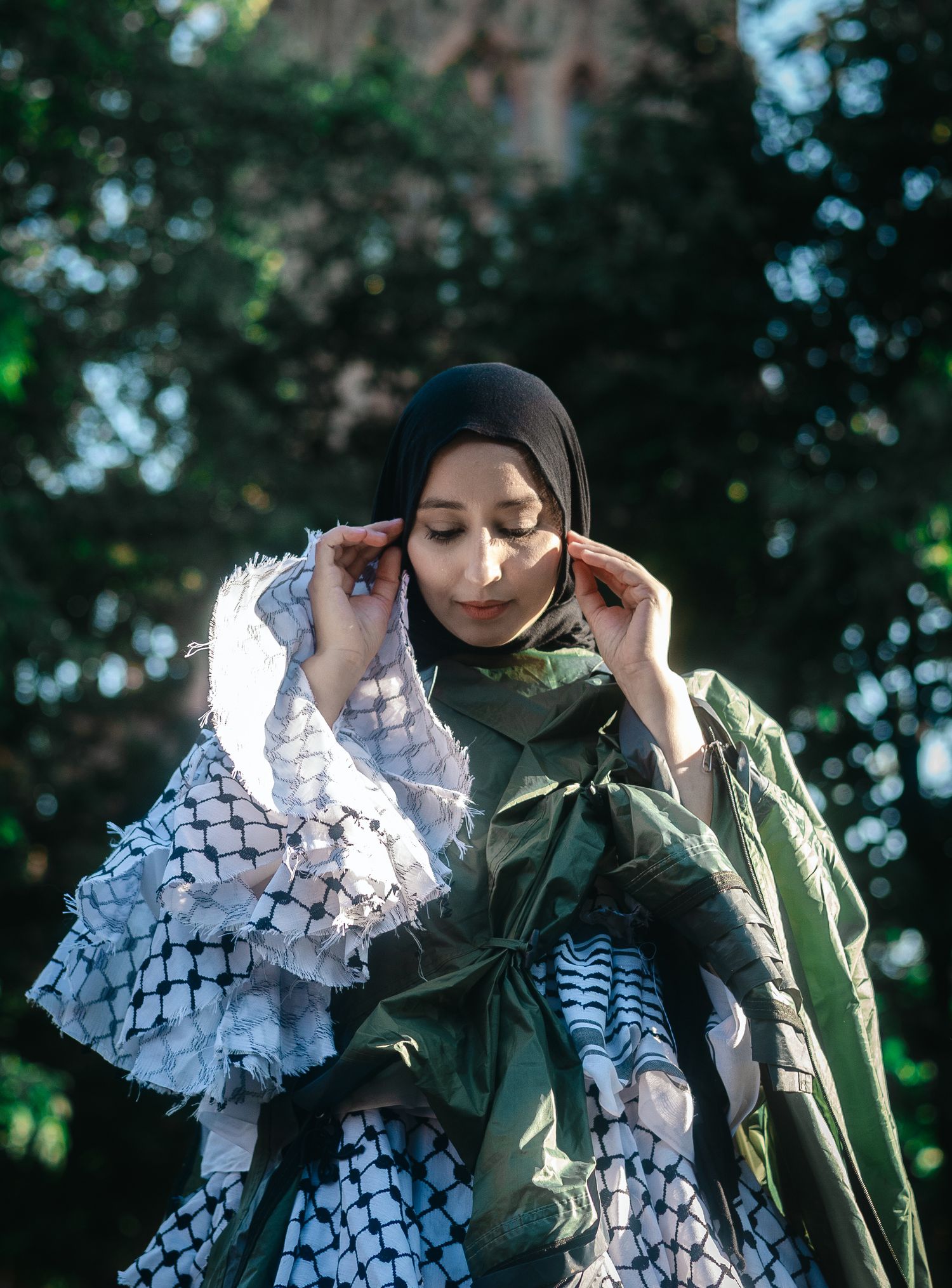 Person wearing a white patterned scarf and green garment adjusts their headwear in an outdoor setting with trees.