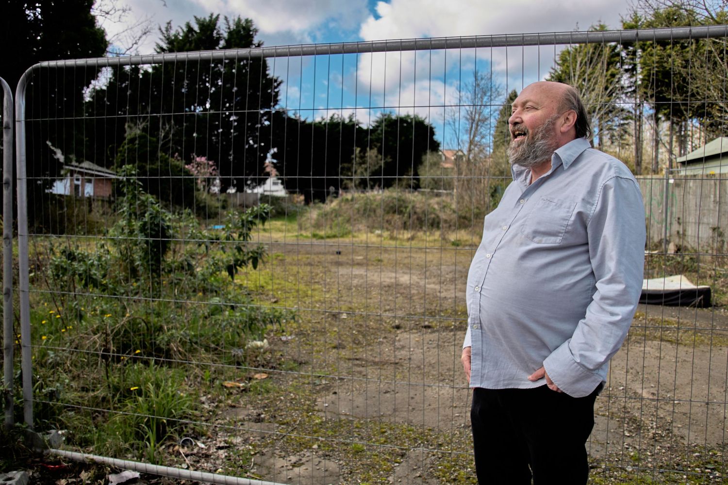 A person observes an empty lot through metal fencing on a sunny day.