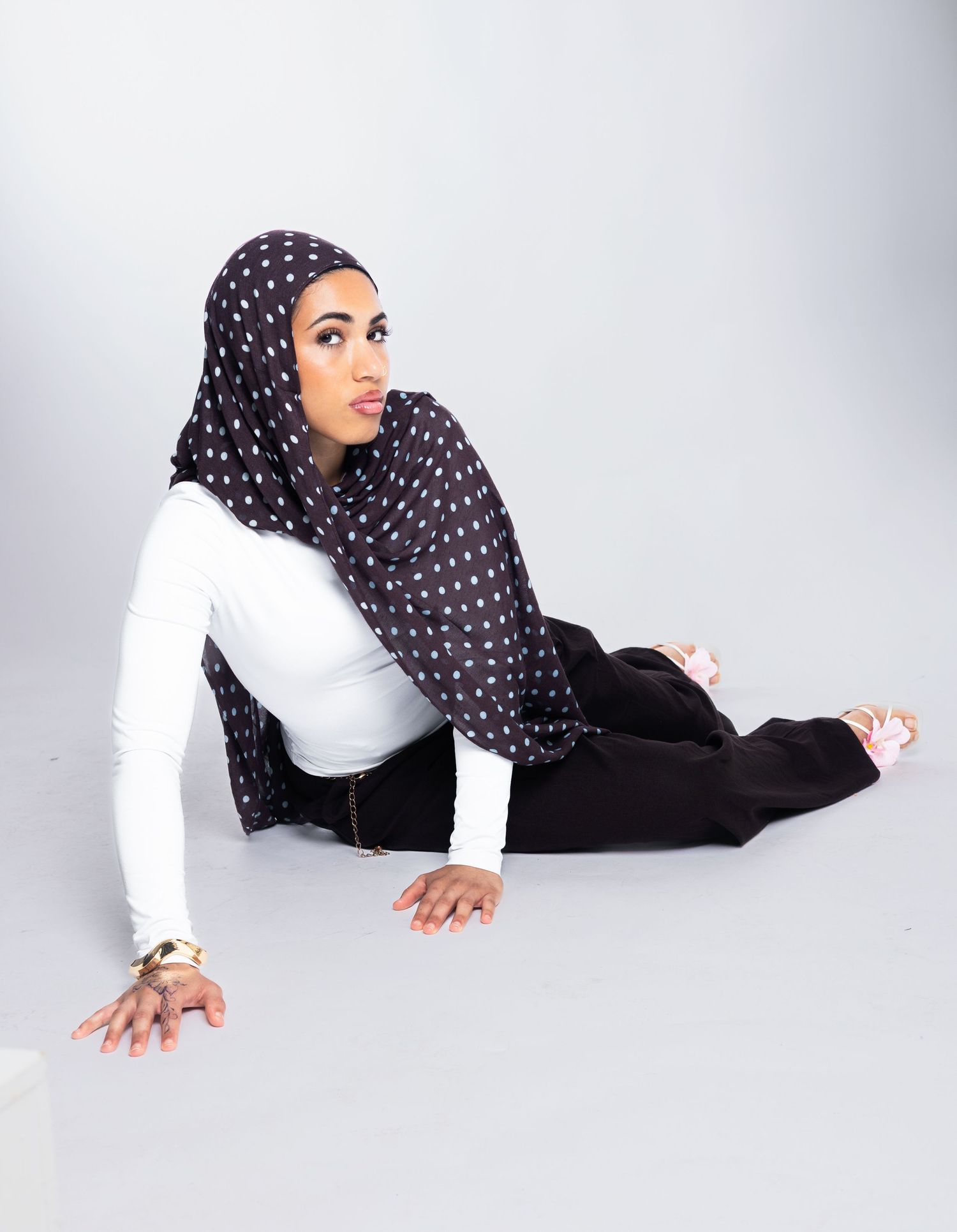 Woman in white top, black pants, and polka dot hijab posing gracefully on the floor in a studio setting.