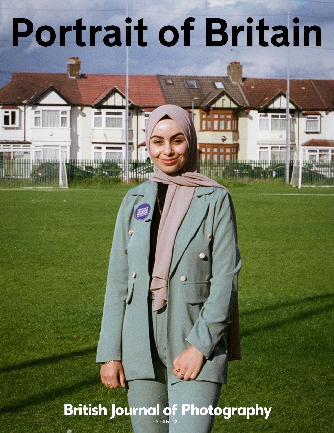 A figure wearing a mint green blazer stands on a grassy field with residential houses visible in the background.