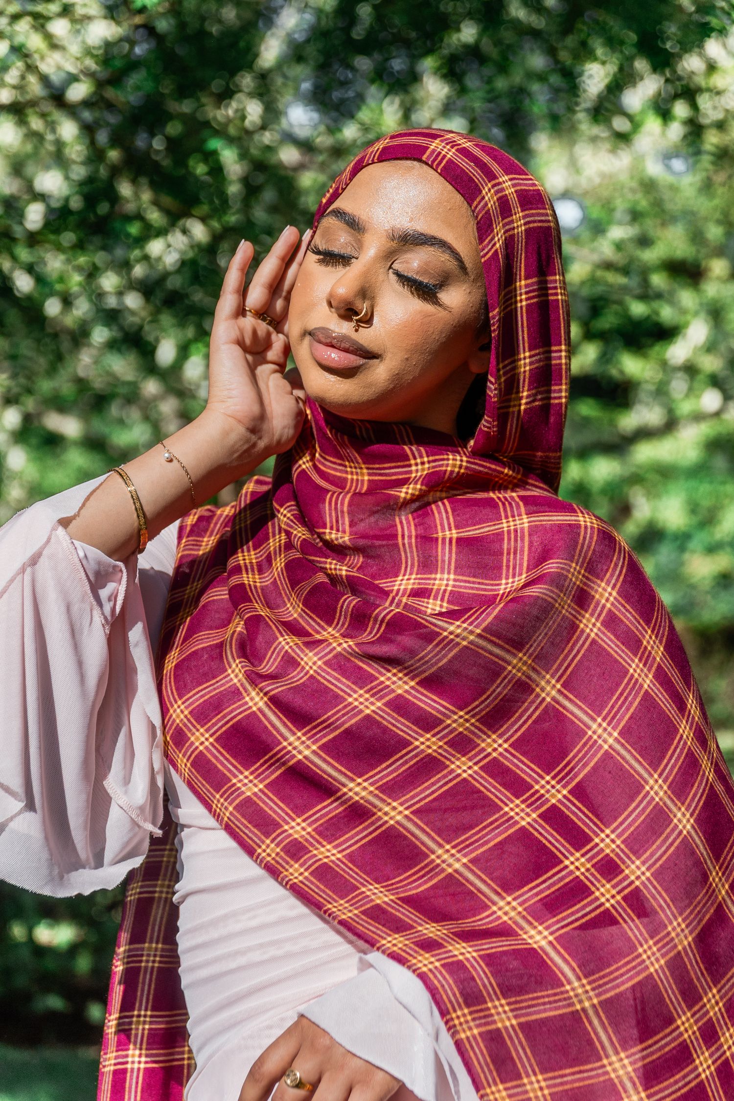 Woman wearing a burgundy plaid hijab with gold accents poses gracefully outdoors with eyes closed.